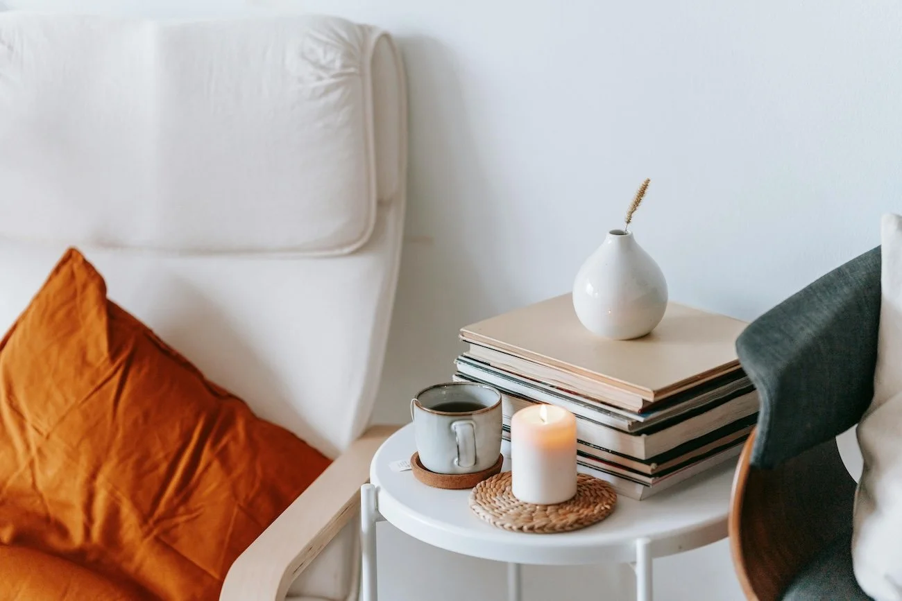 A white round side table next to a bed with a white headboard, an orange pillow, and a green pillow. On the table are a stack of books, a small white vase with a dry plant, a ceramic cup of coffee or tea on a coaster, and a lit candle on a woven coaster.