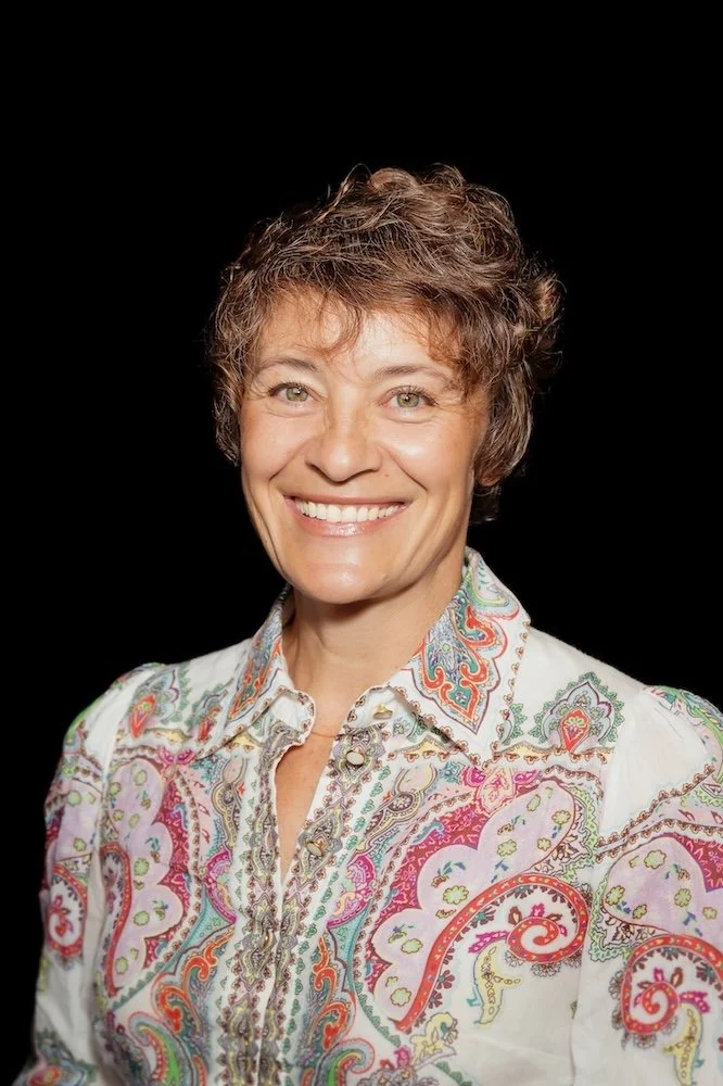 Smiling woman with short curly hair wearing a colorful paisley-patterned blouse against a black background.