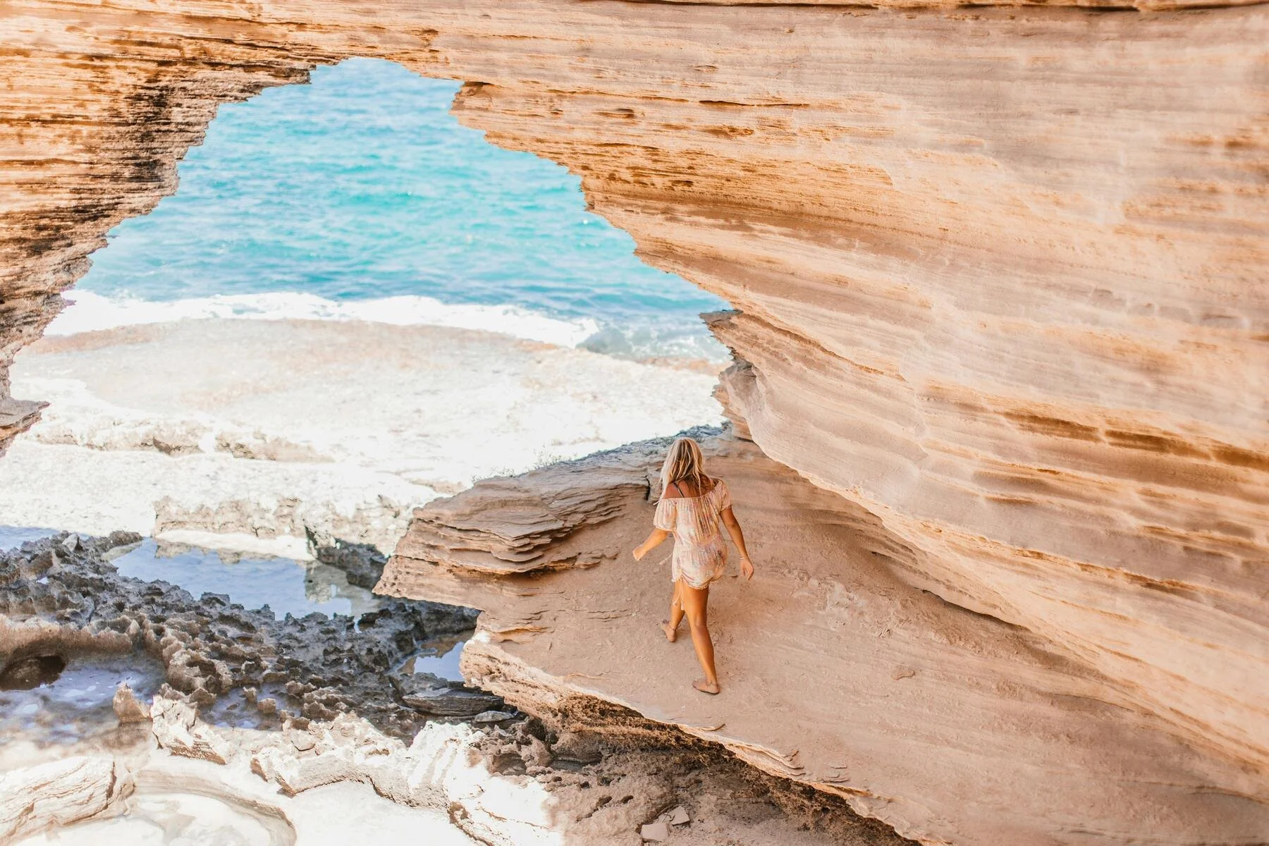 A woman walking on a sandy beach through a natural rock archway with the ocean and sky visible in the background. A representation of the approach naturopath Eloise smith from clean slate naturopathy takes