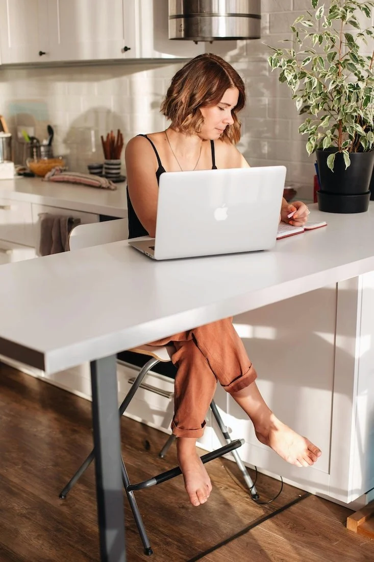 A woman sitting at a kitchen island working on a laptop with her bare feet slightly off the ground and a notebook nearby, in a modern kitchen with a large plant on the counter.