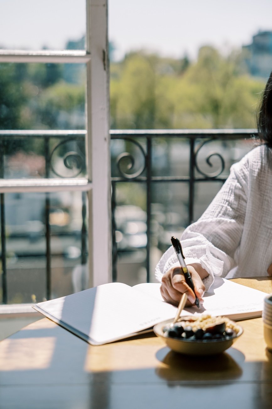 Naturopath in bayside melbourne showing a Person sitting at a table, writing in a notebook with a pen, near an open window with a scenic outdoor view.