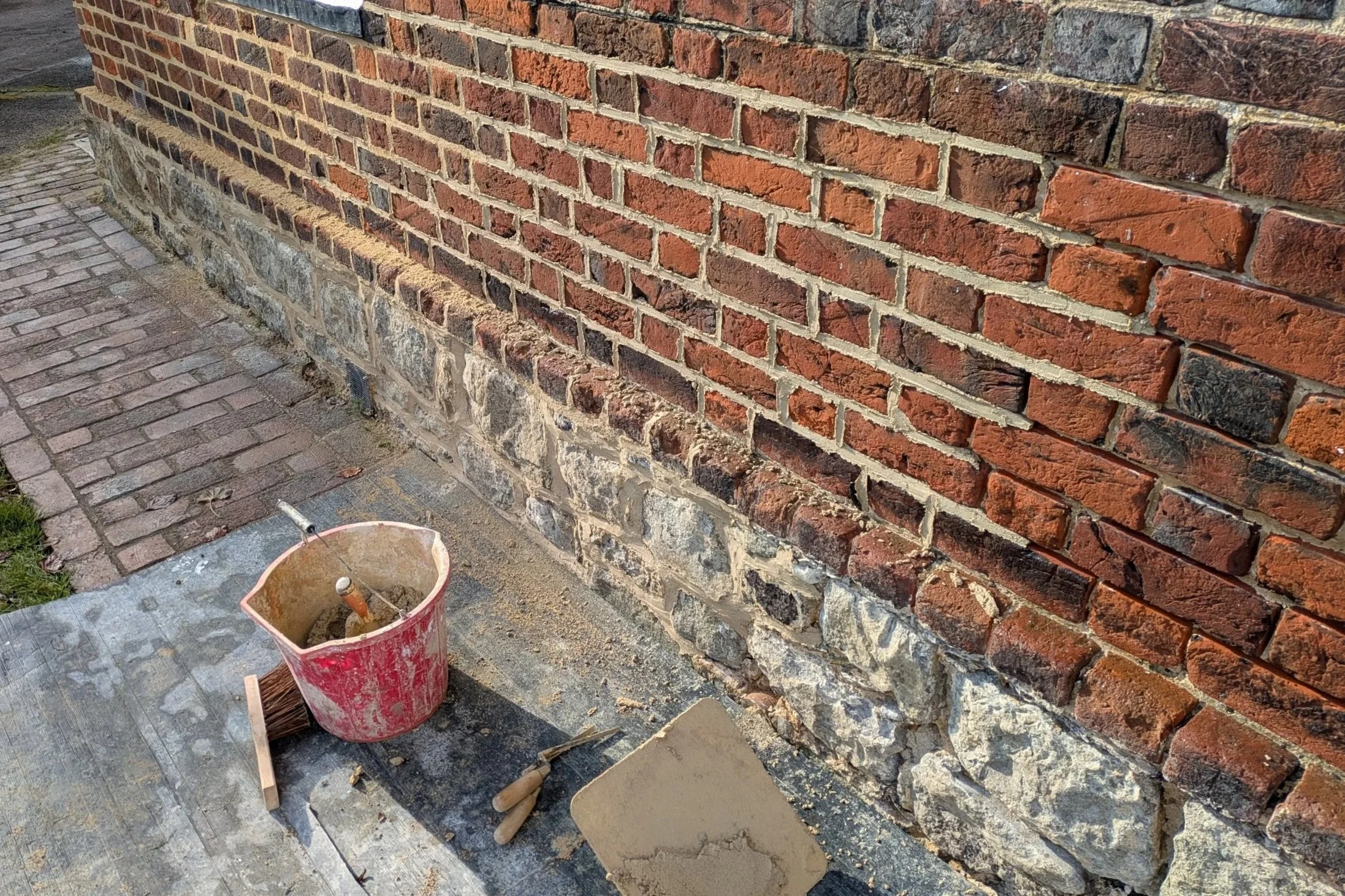 A brick wall being restored using lime mortar, with a mixture of red bricks and stones at the base. There is a bucket with masons' tools, including a trowel, hawk and a brush.