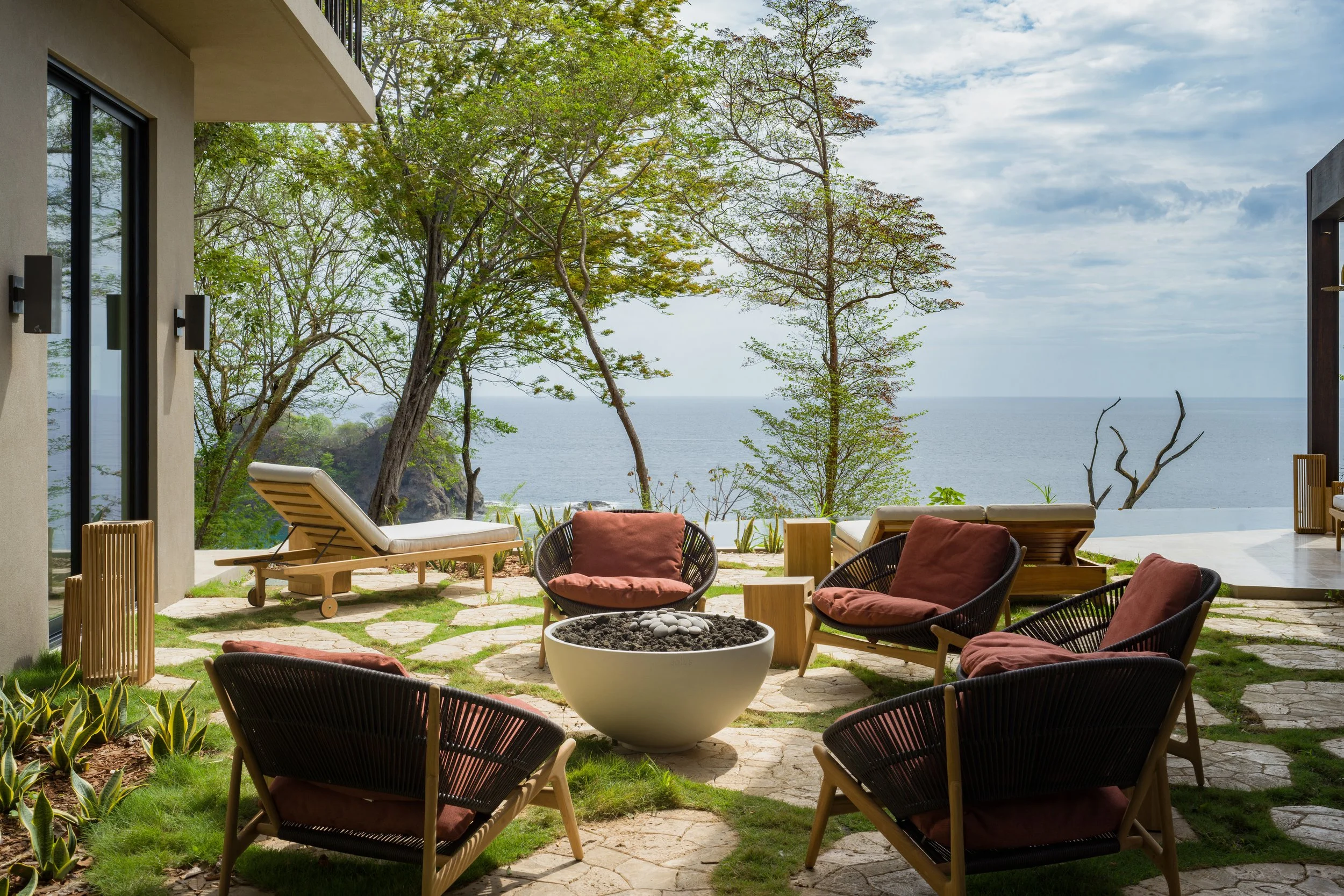 Outdoor patio area with six chairs with cushions surrounding a fire pit, a lounge chair, and a view of the ocean framed by trees and a partly cloudy sky.