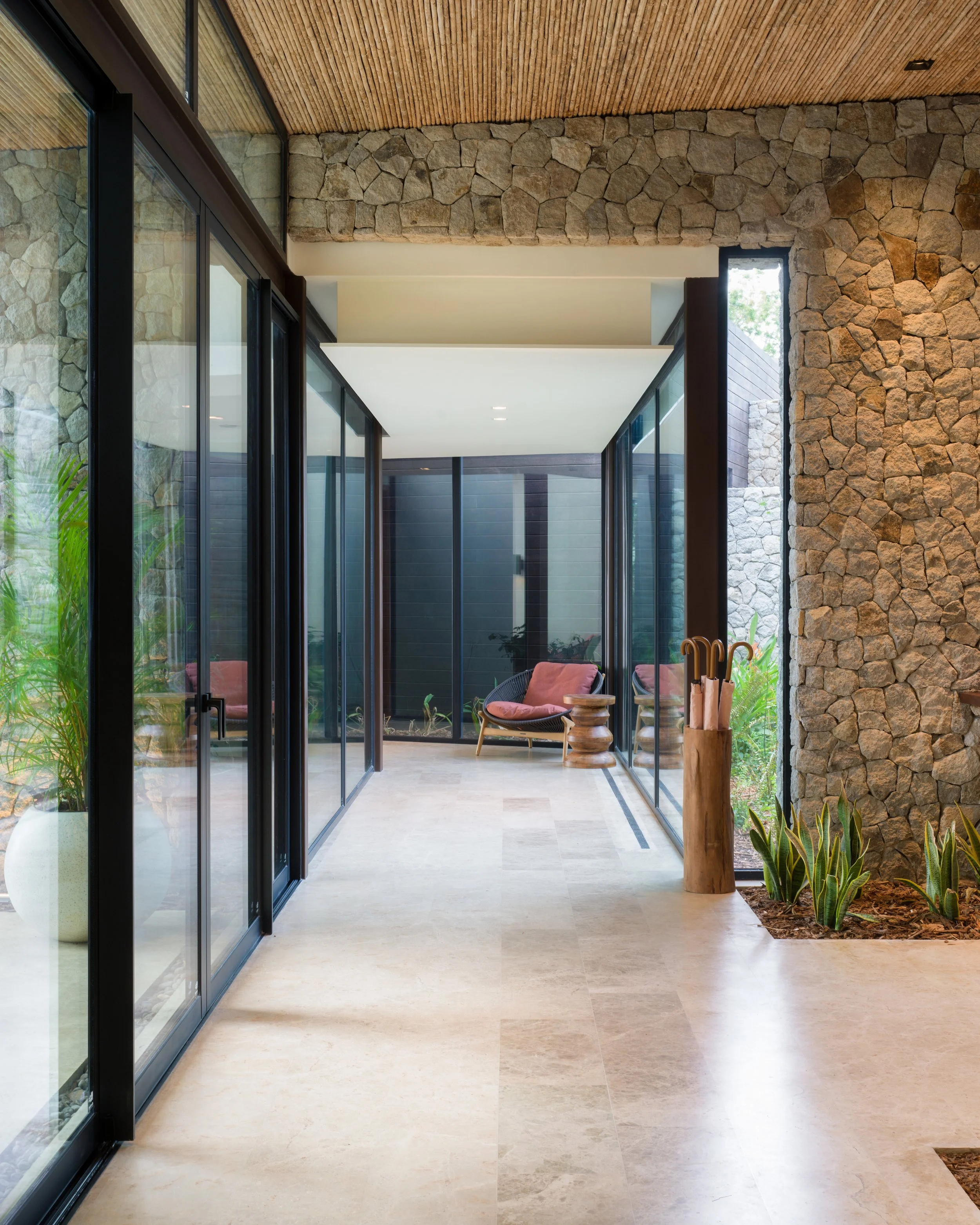 Sunlit indoor corridor with glass sliding doors, stone and wood interior, potted plants, and seating area with pink cushions.