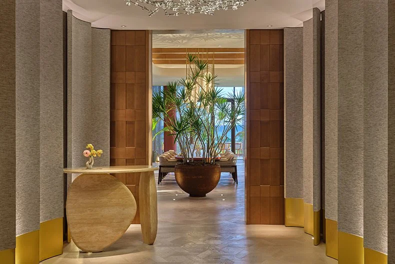 Elegant hotel lobby with a large potted plant in the center, wooden accents, and a seating area in the background near large windows.