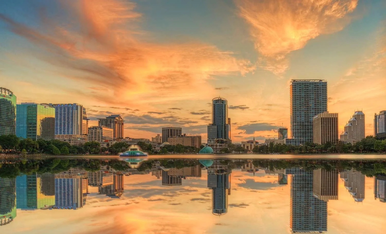 Sunset view of a city skyline with skyscrapers, reflected in a body of water with a fountain, trees, and partly cloudy sky.