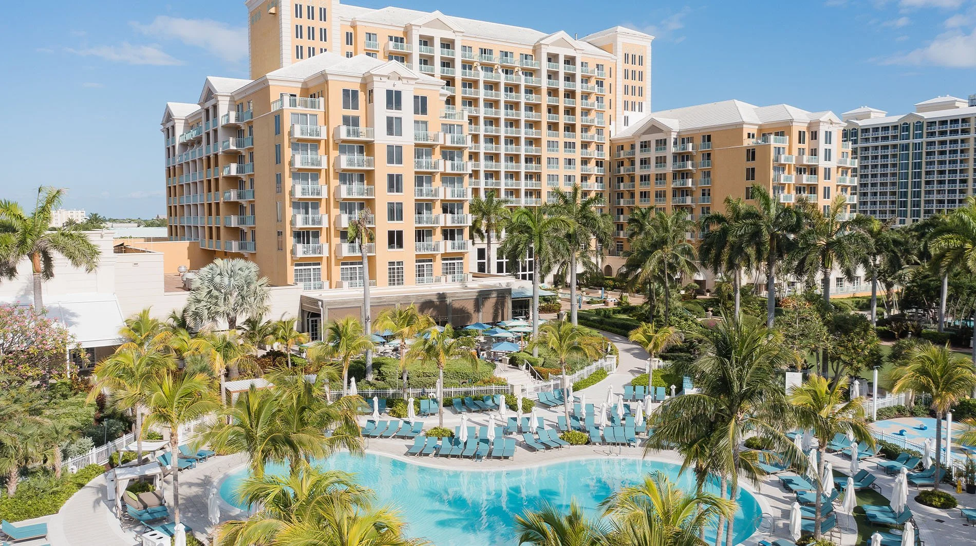A large beige-colored hotel with multiple balconies, surrounded by lush tropical plants and tall palm trees. In the foreground, there is a swimming pool with lounge chairs and umbrellas, set in a well-maintained garden area on a sunny day.