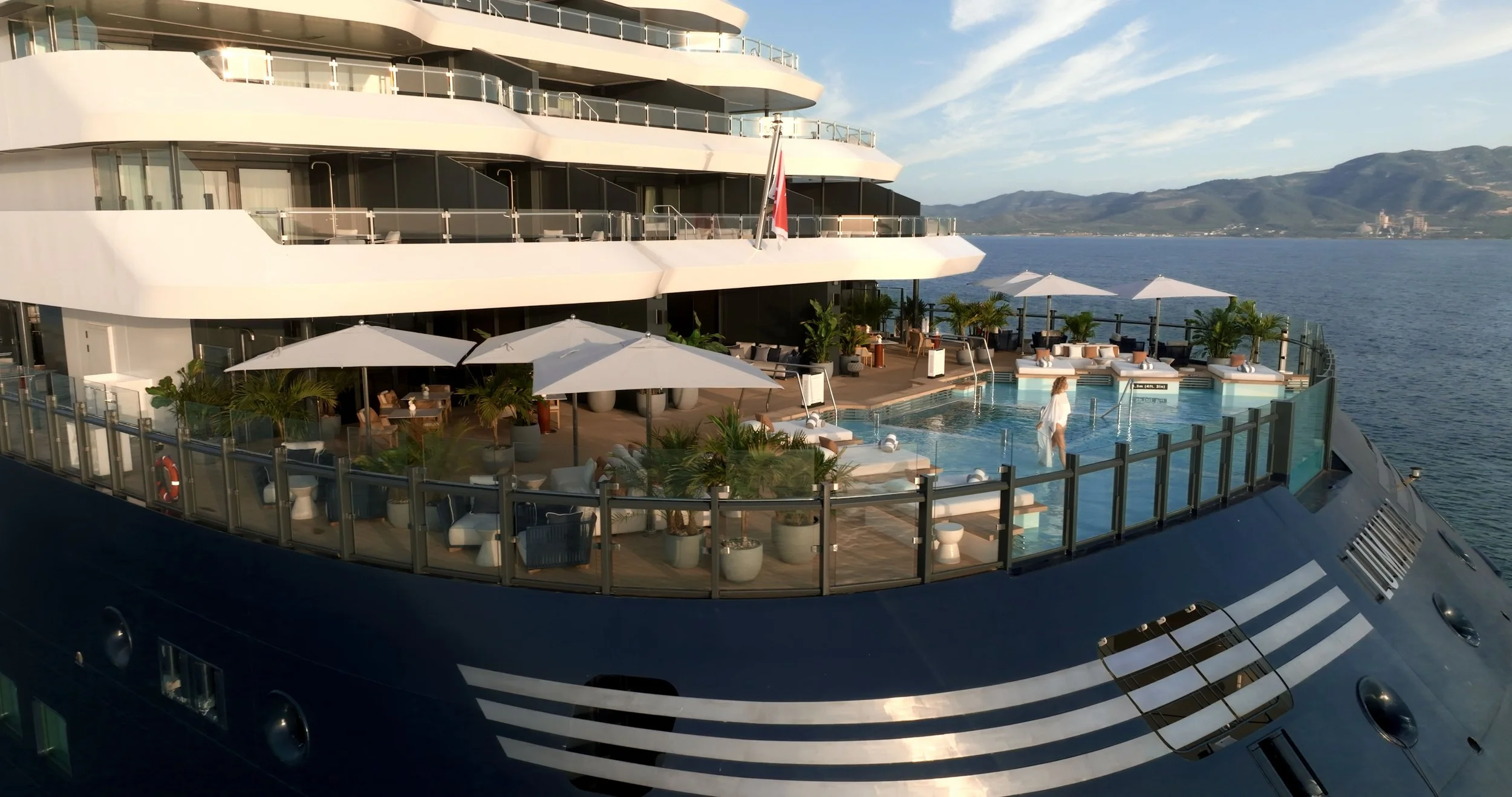 A luxury yacht with multiple decks, an outdoor pool area, and lounge chairs. The yacht is on the water with a scenic mountainous landscape in the background.