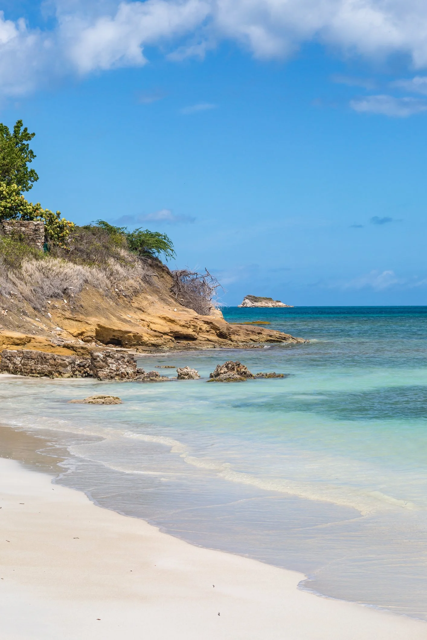 A scenic view of a beach with sandy shore, rocks in the shallow water, a rocky cliff with trees, and a small island in the distance under a partly cloudy sky.