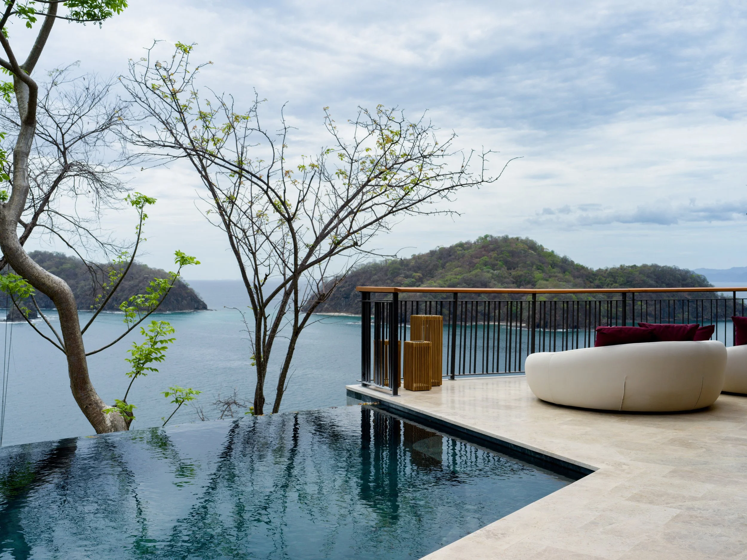 Luxe outdoor pool area overlooking a coastal ocean with hills in the background, featuring modern white seating and wooden accents, under a partly cloudy sky.