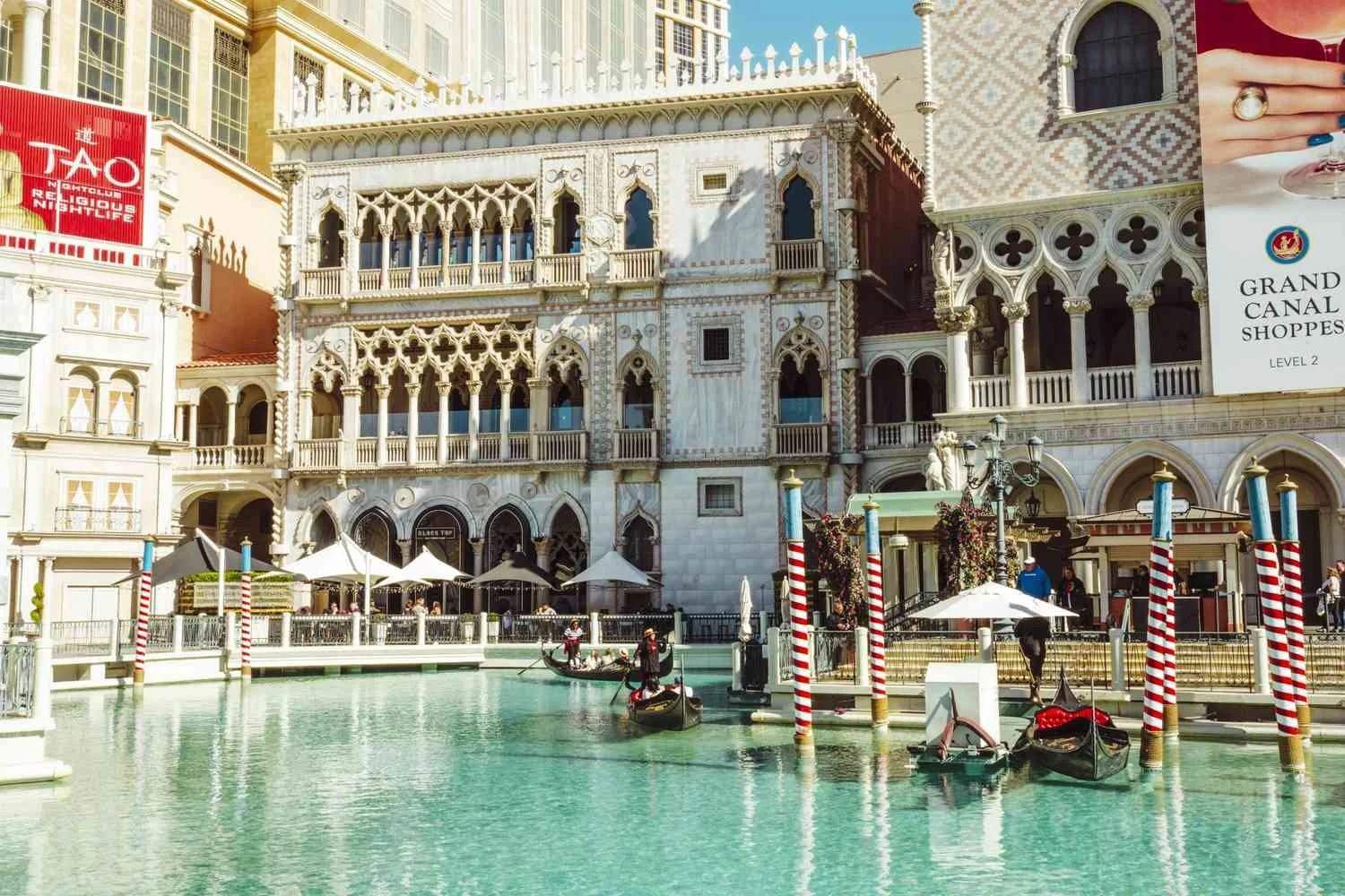 Replica of Venice's Grand Canal with gondolas, small cafes with umbrellas, and a backdrop resembling Venice's architecture, in a themed shopping complex.