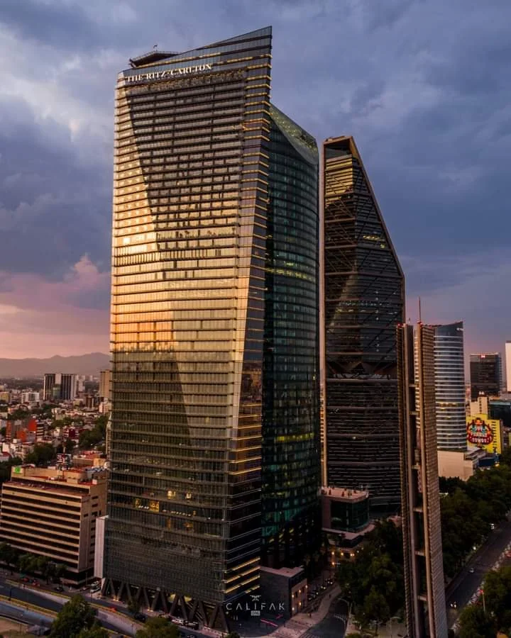 Tall modern glass skyscrapers in a cityscape at sunset, with one building labeled 'The Trump Tower' and a mix of other high-rise buildings and a distant mountain range in the background.
