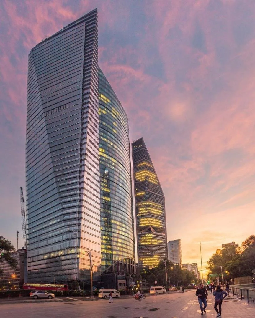 Tall modern skyscrapers with glass facades at sunset, reflecting pink and purple sky, busy city street with cars and pedestrians.