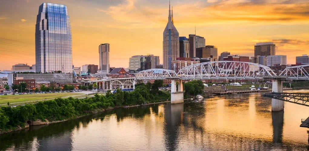 City skyline of Nashville Tennessee at sunset, featuring skyscrapers, a bridge over the river, and a colorful sky.