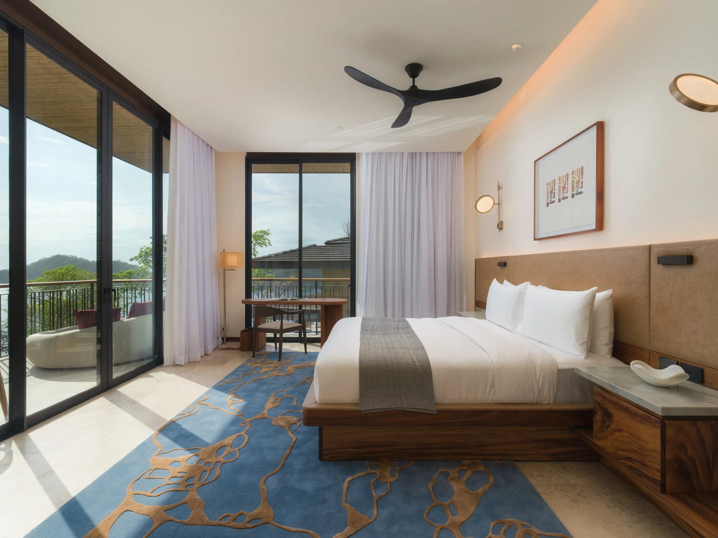 Modern hotel room with large glass sliding doors, a balcony with a lounge chair, and an interior featuring a king-sized bed with white bedding, a wooden nightstand, a blue patterned rug, and wall-mounted lamps.