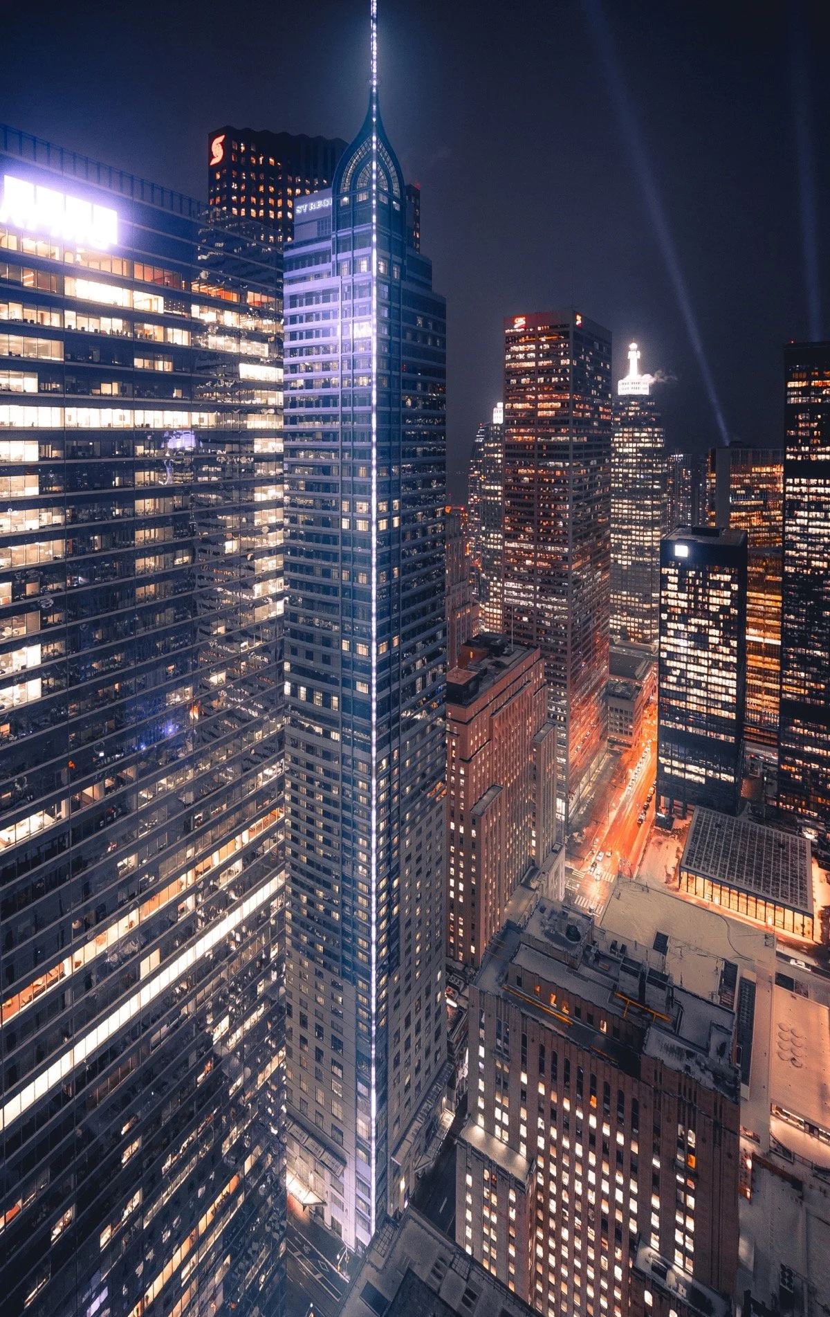 Nighttime cityscape of illuminated skyscrapers in a downtown area, with streets below and lights on in the buildings.