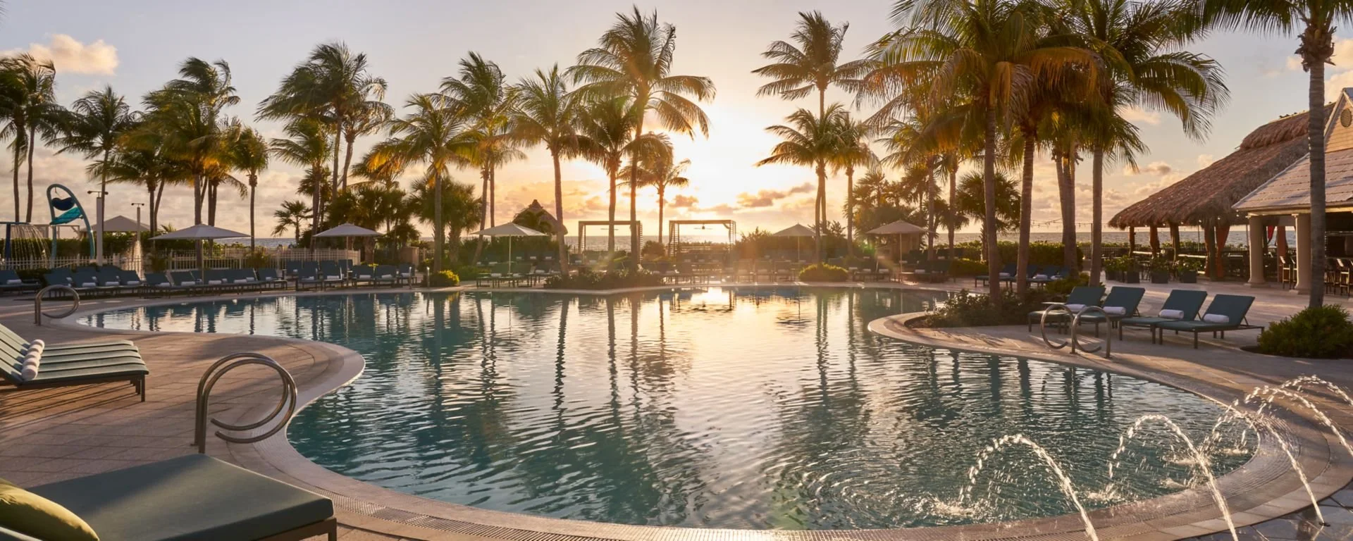 A tropical resort swimming pool scene at sunset with palm trees, lounge chairs, umbrellas, and a thatched-roof tiki bar.