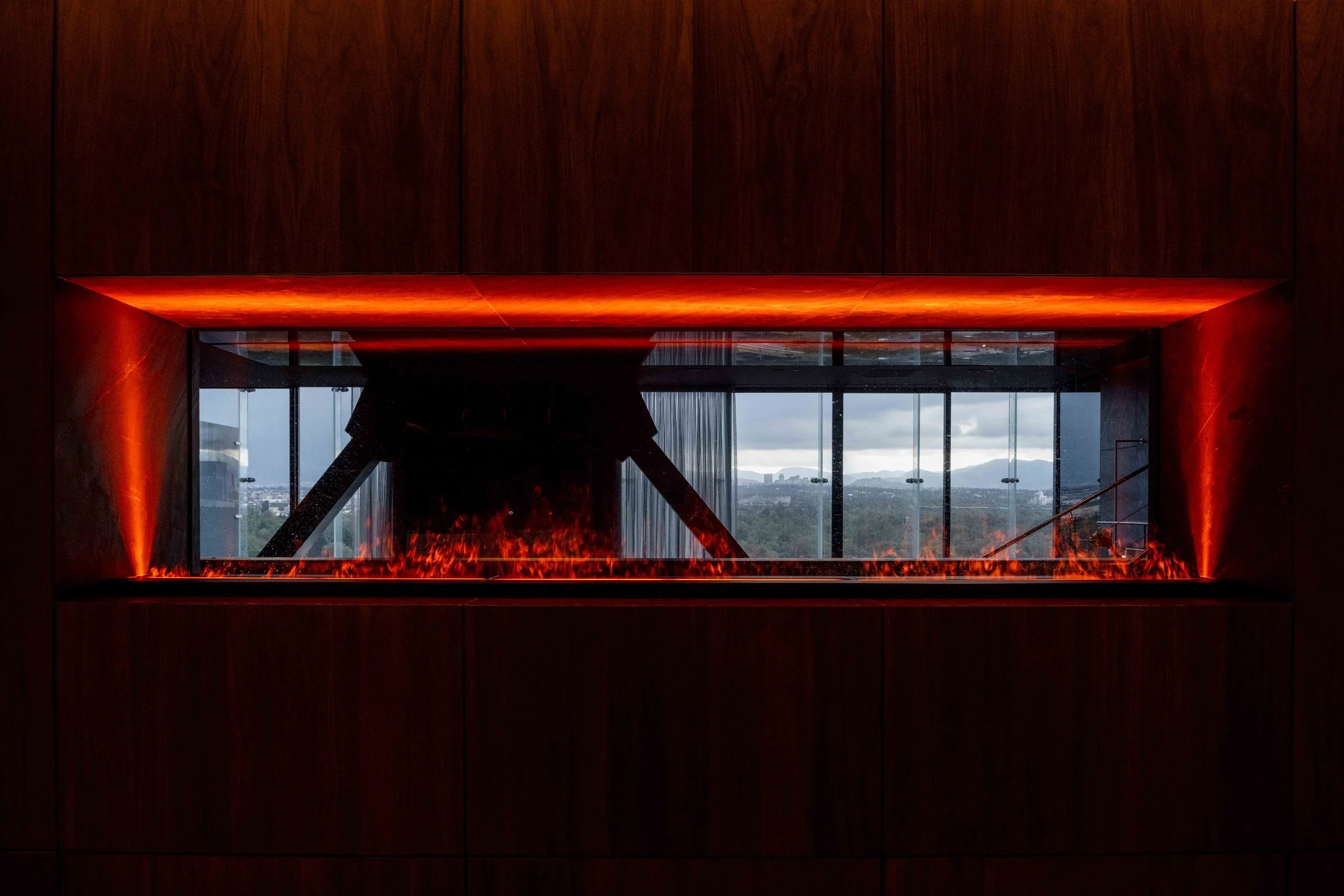 Interior view of a modern room with a rectangular fireplace, glowing with orange and red flames, surrounded by dark wood paneling, and large glass windows showing an outdoor landscape with hills and cloudy sky.