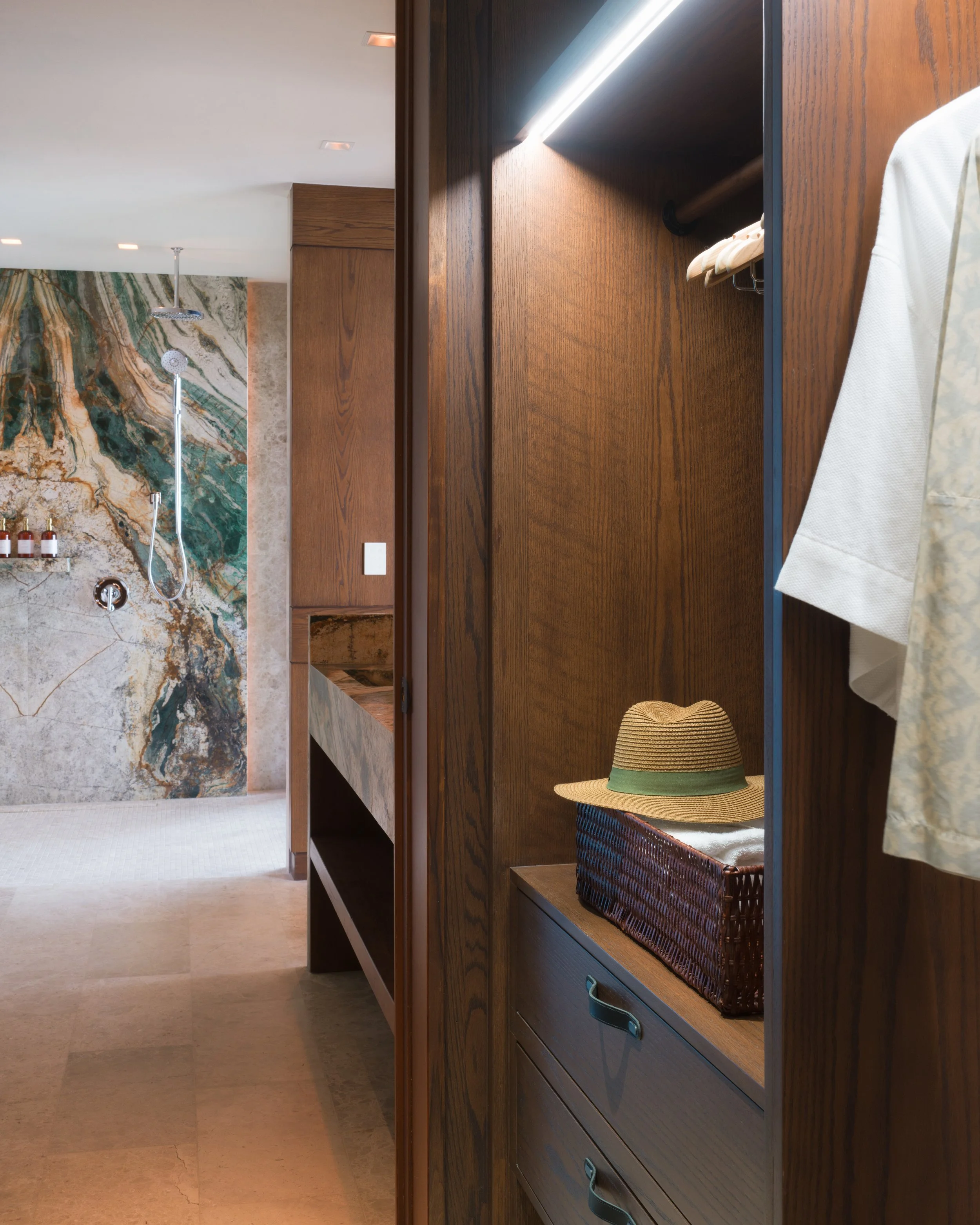 Open wardrobe with a straw hat and clothing inside, view of a bathroom with a marble shower wall and rainfall shower head.