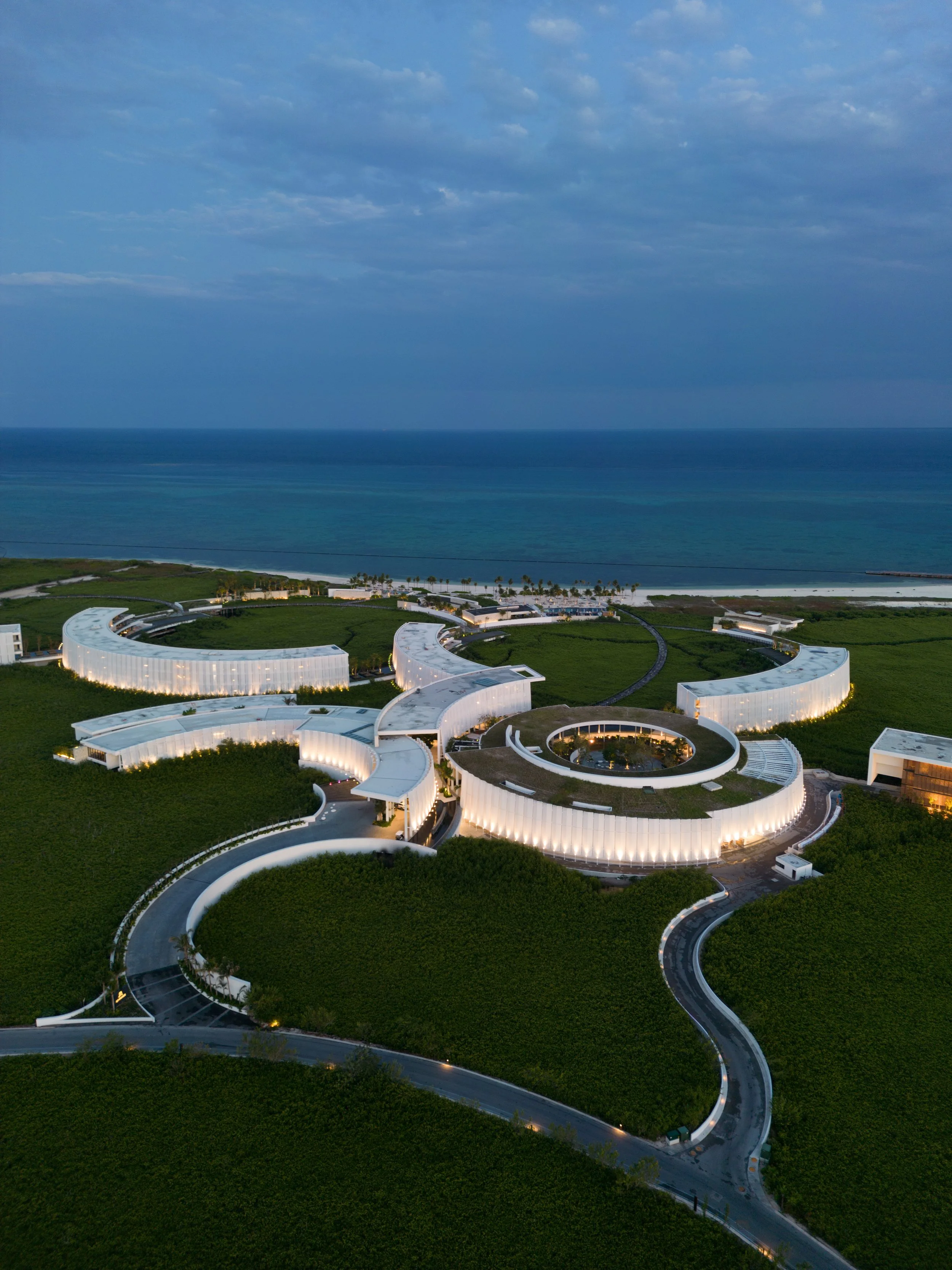 Aerial view of a modern, curved white hotel or resort building complex near the beach, with green landscaping and winding roads around it, overlooking the ocean under a cloudy sky.