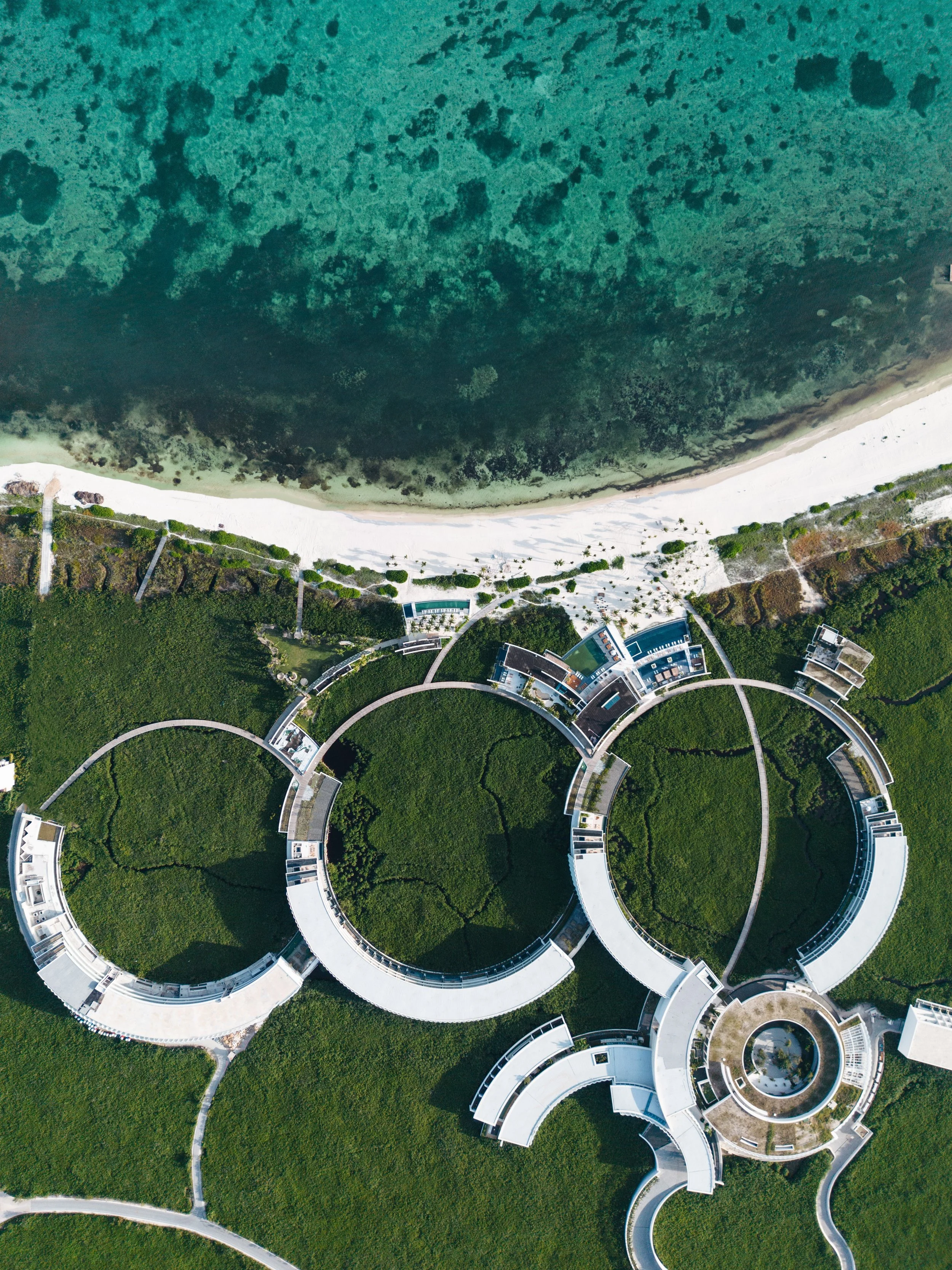 Aerial view of modern circular buildings surrounded by green landscape, with a beach and ocean in the background.