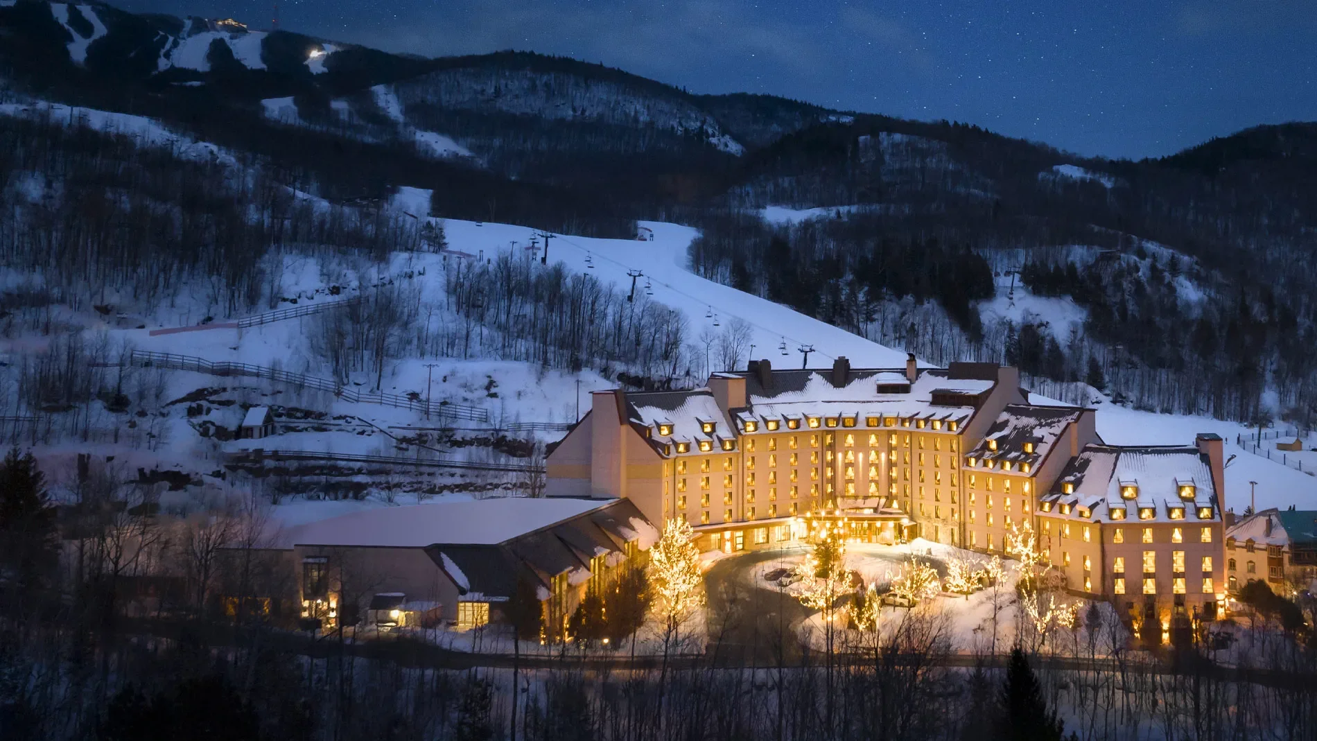 A nighttime view of a large, multi-story hotel with lit windows and decorative outdoor lights, surrounded by snow-covered trees and mountains with ski trails, under a starry sky.