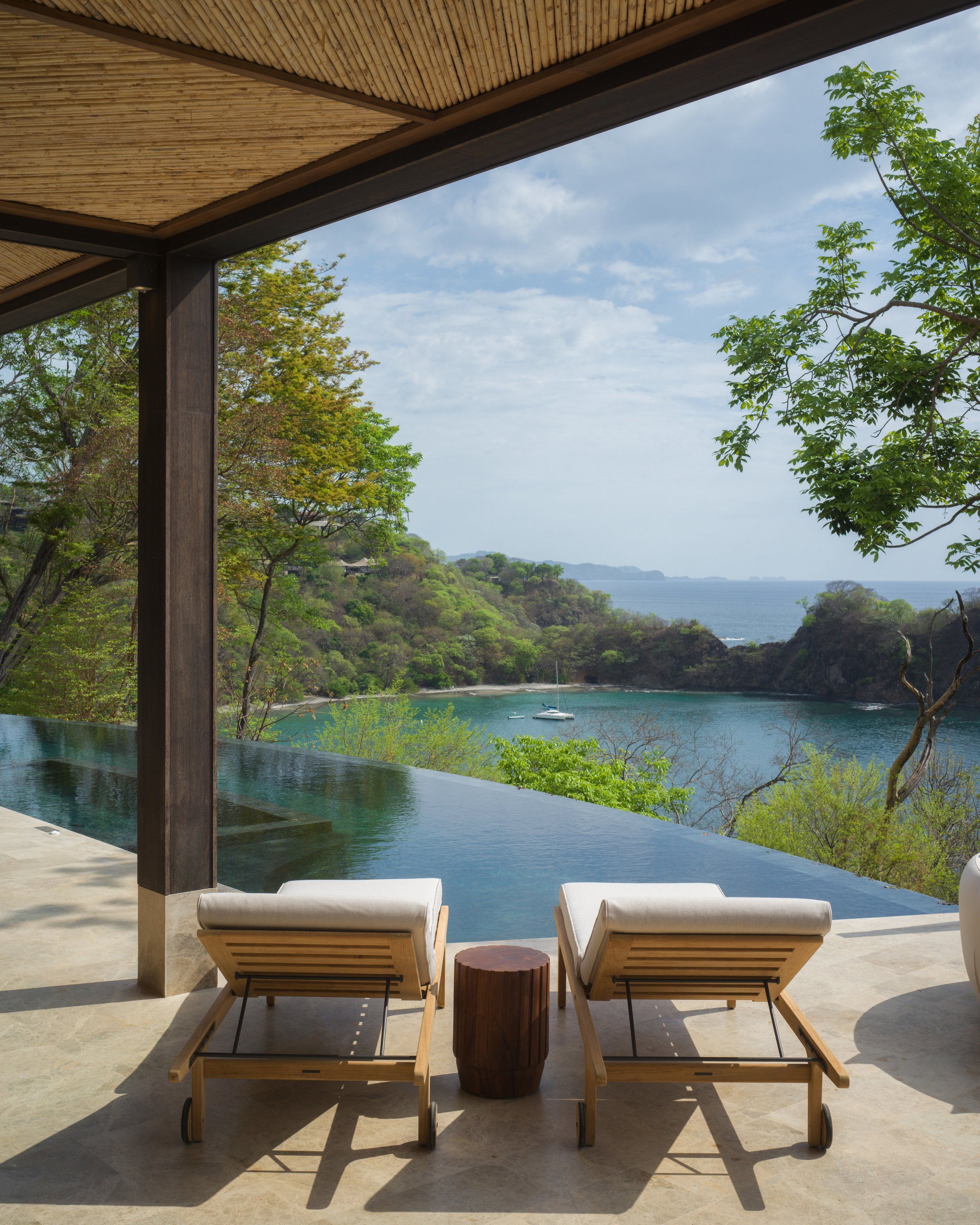 Two lounge chairs on a patio facing an infinity pool with a view of a bay, green trees, and a sailboat in the water, under a wooden roof.