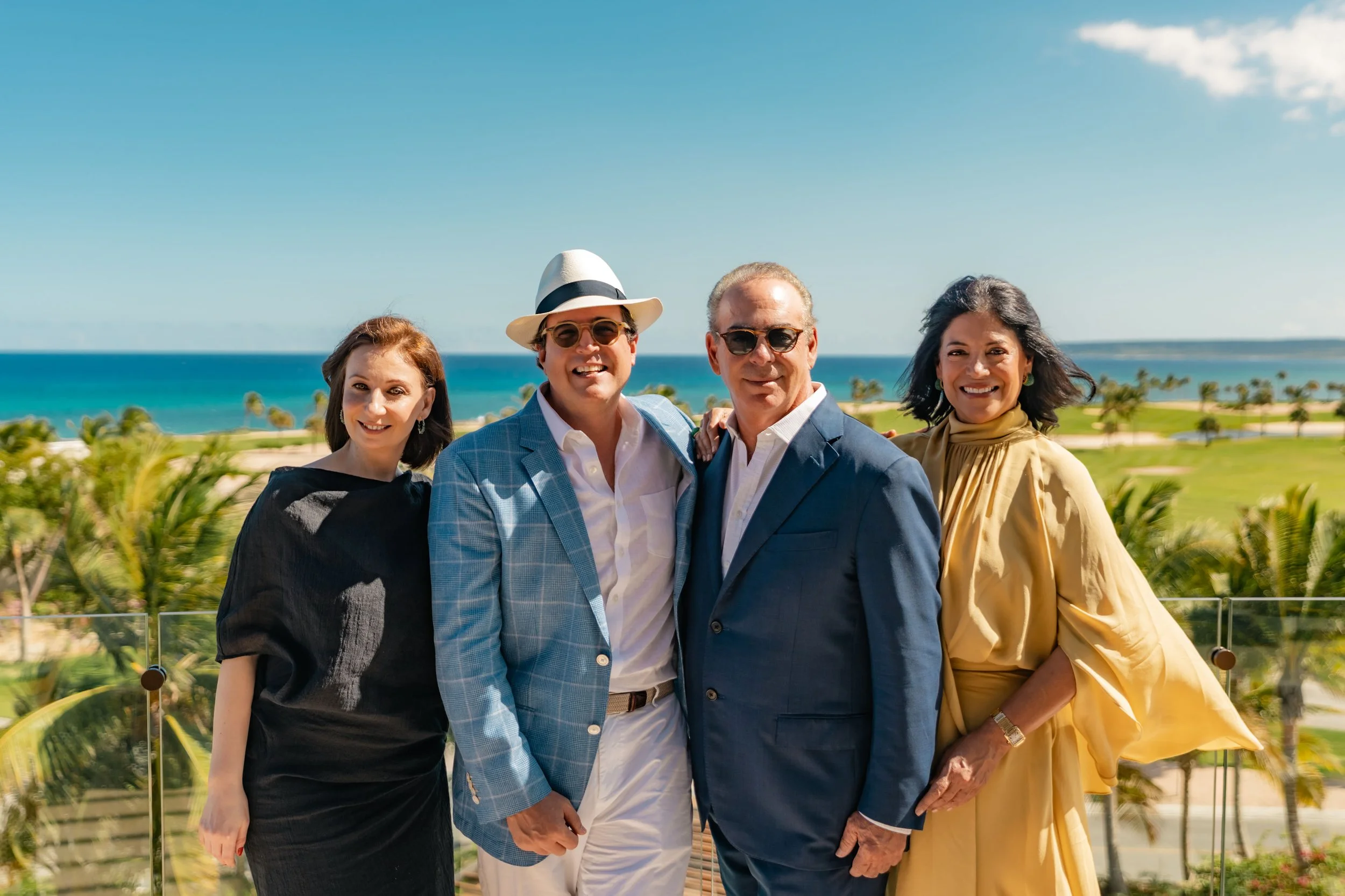 Group of four people standing outdoors on a balcony with ocean in the background, smiling at the camera.