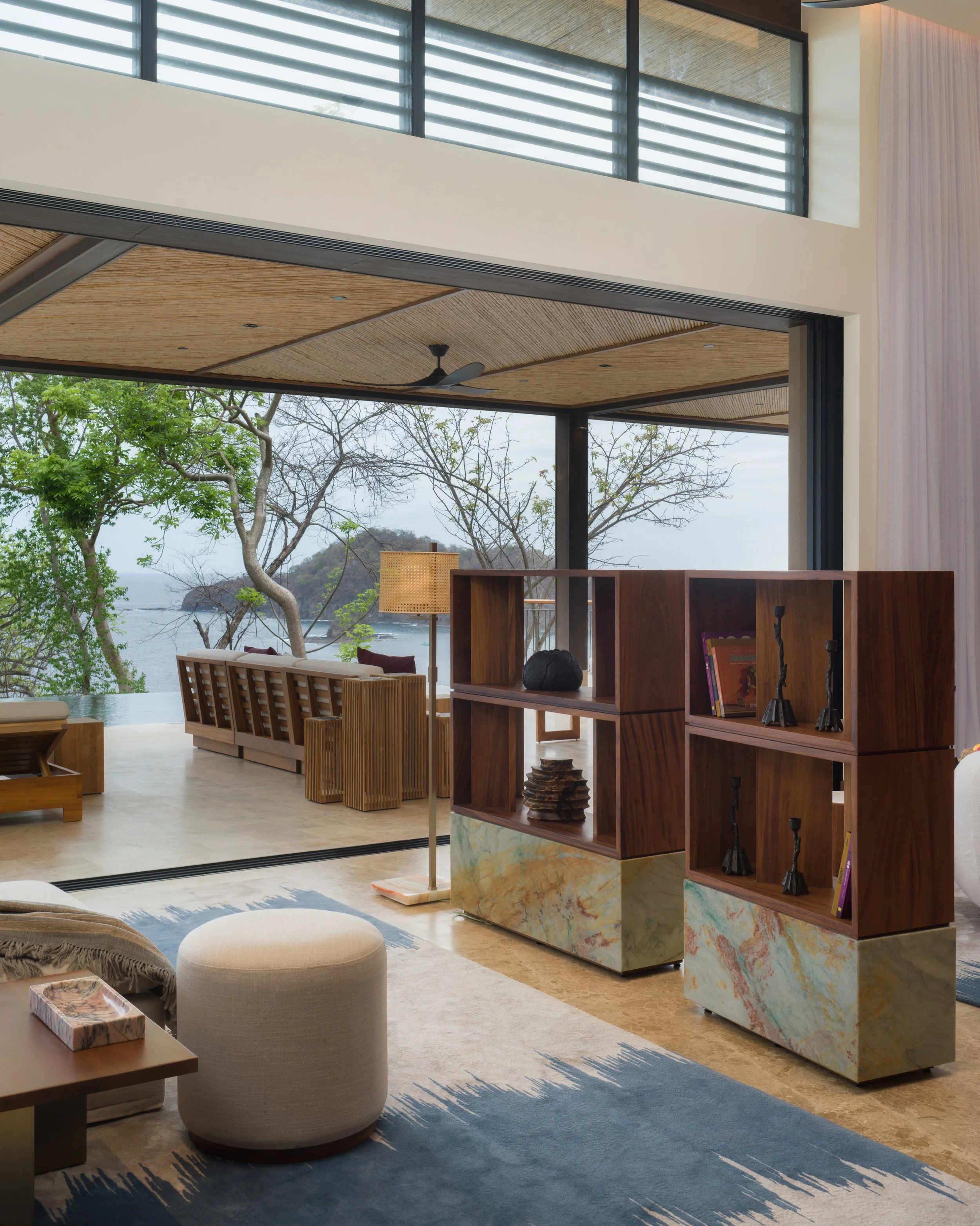 Living room with a large open window overlooking trees and a body of water, with modern decor including a beige pouf, wooden shelving, and a blue area rug.
