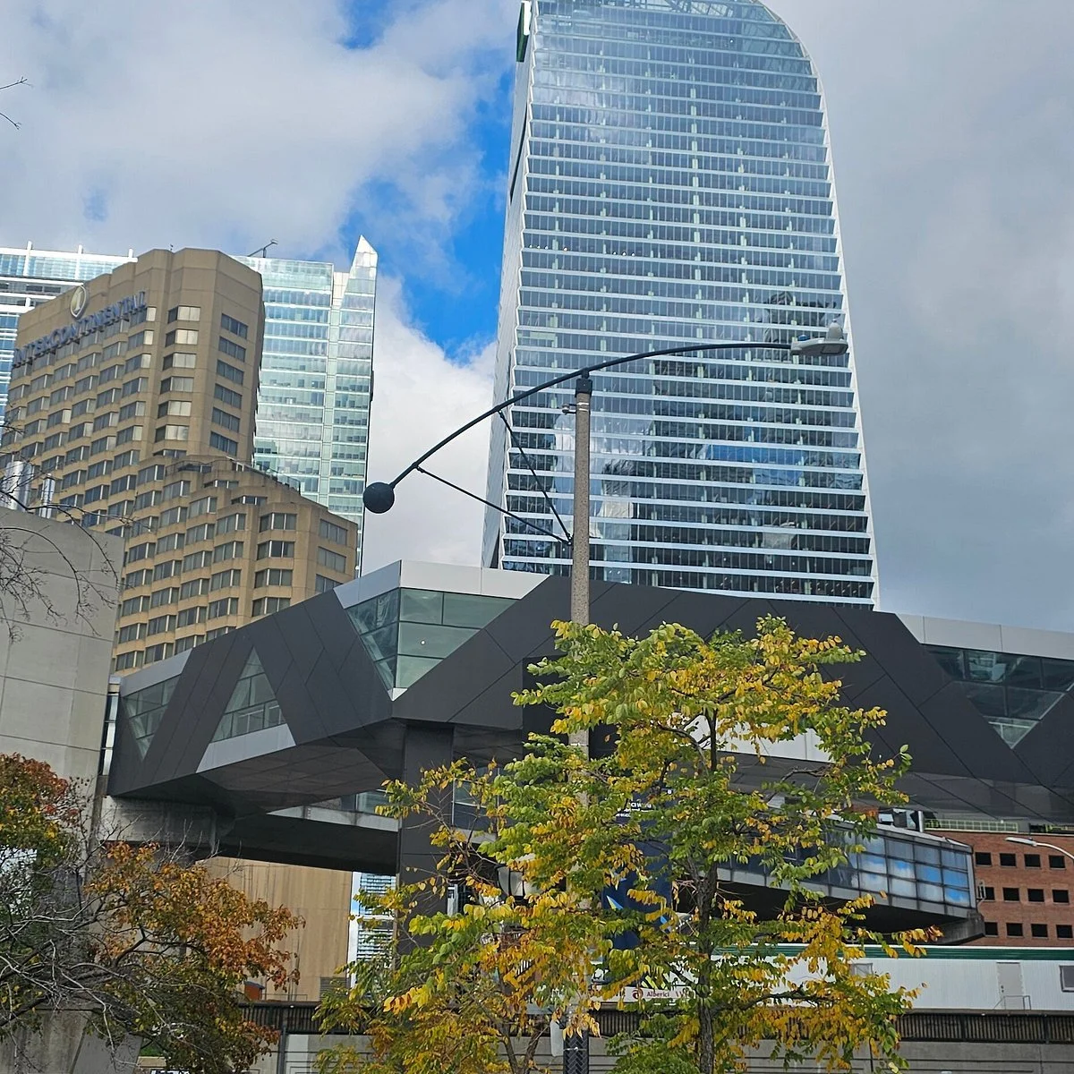 Tall modern skyscrapers with glass facades in a city, with a curved skybridge connecting buildings, streetlamp in foreground, and a tree with yellow and green leaves.