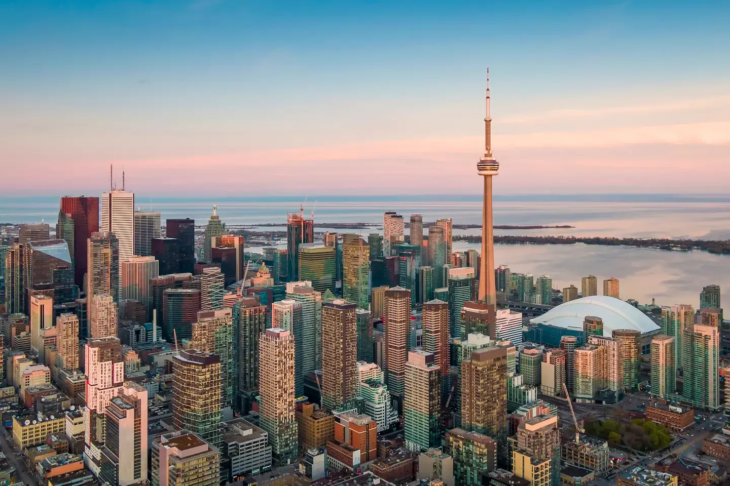 Skyline of Toronto, Canada, with CN Tower, Rogers Centre stadium, high-rise buildings, and Lake Ontario in the background during sunset.