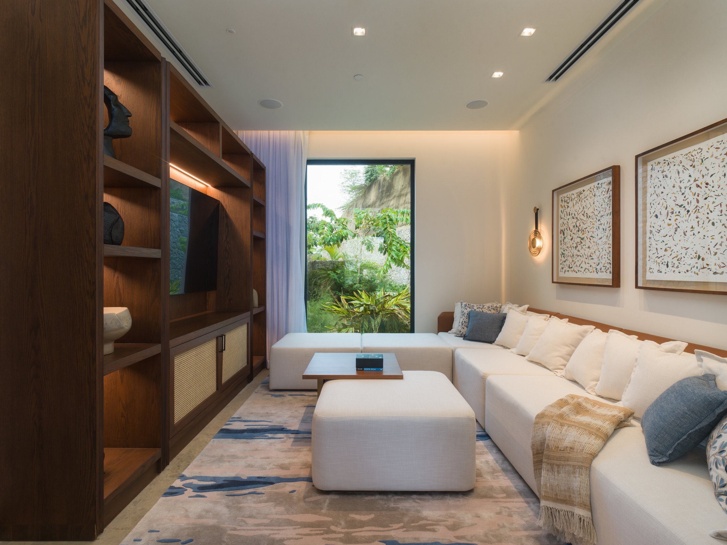 Modern living room with beige sectional sofa, decorative pillows, wooden bookshelf, wall art, and large window showing greenery outside.