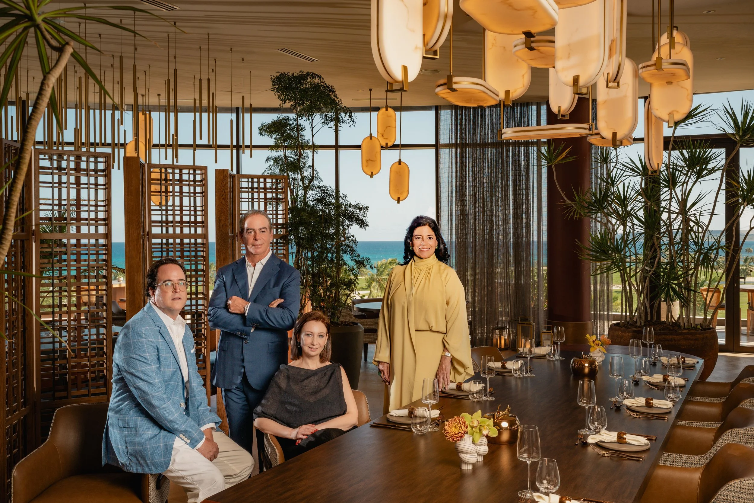 Four people, three men and one woman, in a well-lit, modern dining room with ocean view, decorated with plants and hanging lights, standing around a large dining table with place settings and floral centerpieces.