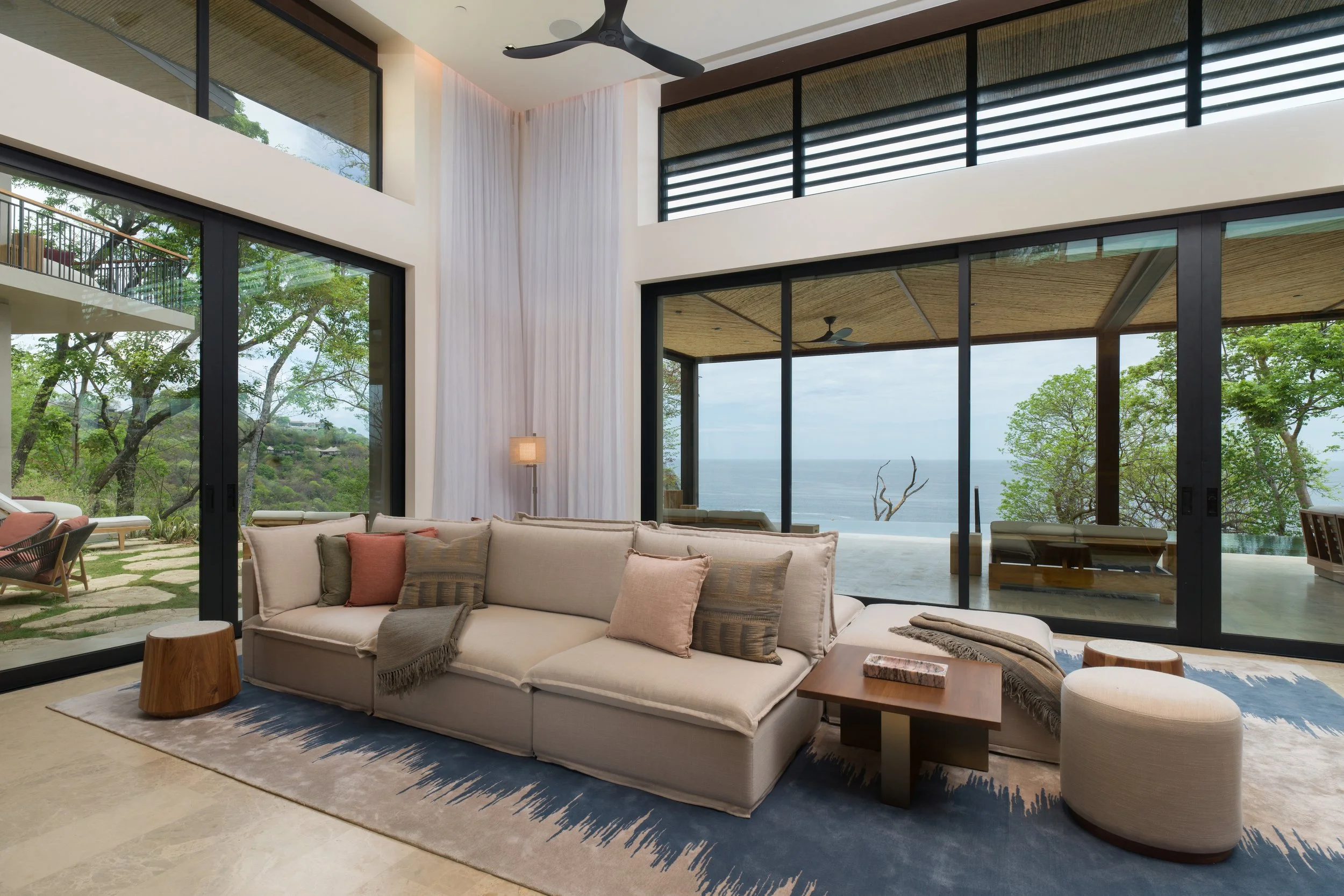 Living room with large floor-to-ceiling glass windows and doors, ocean view, beige sofa with pillows, small wooden coffee table, rug, and outdoor balcony with seating, trees and sky visible outside.