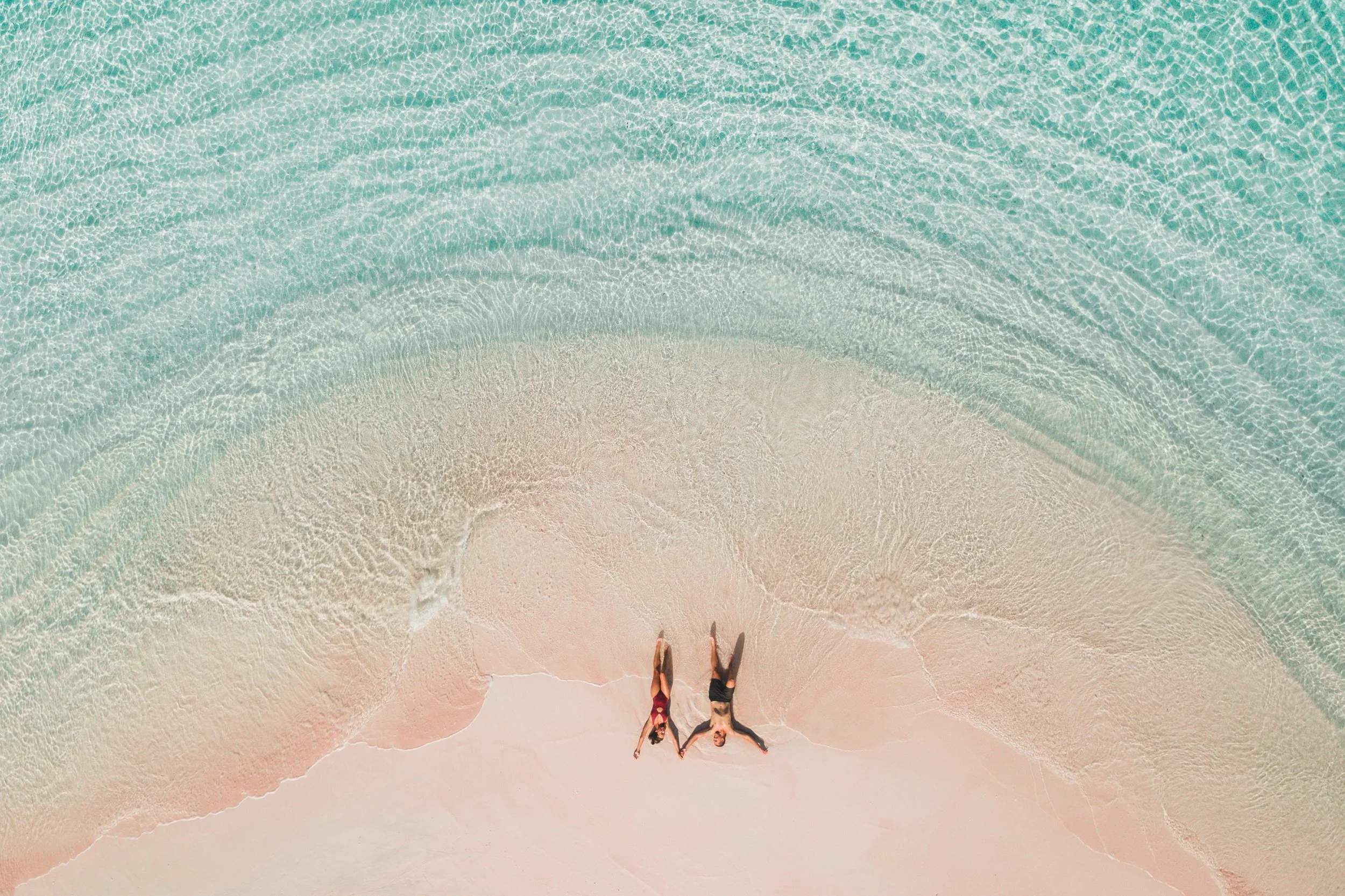 Bird's eye view of a sandy beach with two women lying next to each other, wearing swimsuits, near the water's edge, with clear blue-green ocean waves in the background.