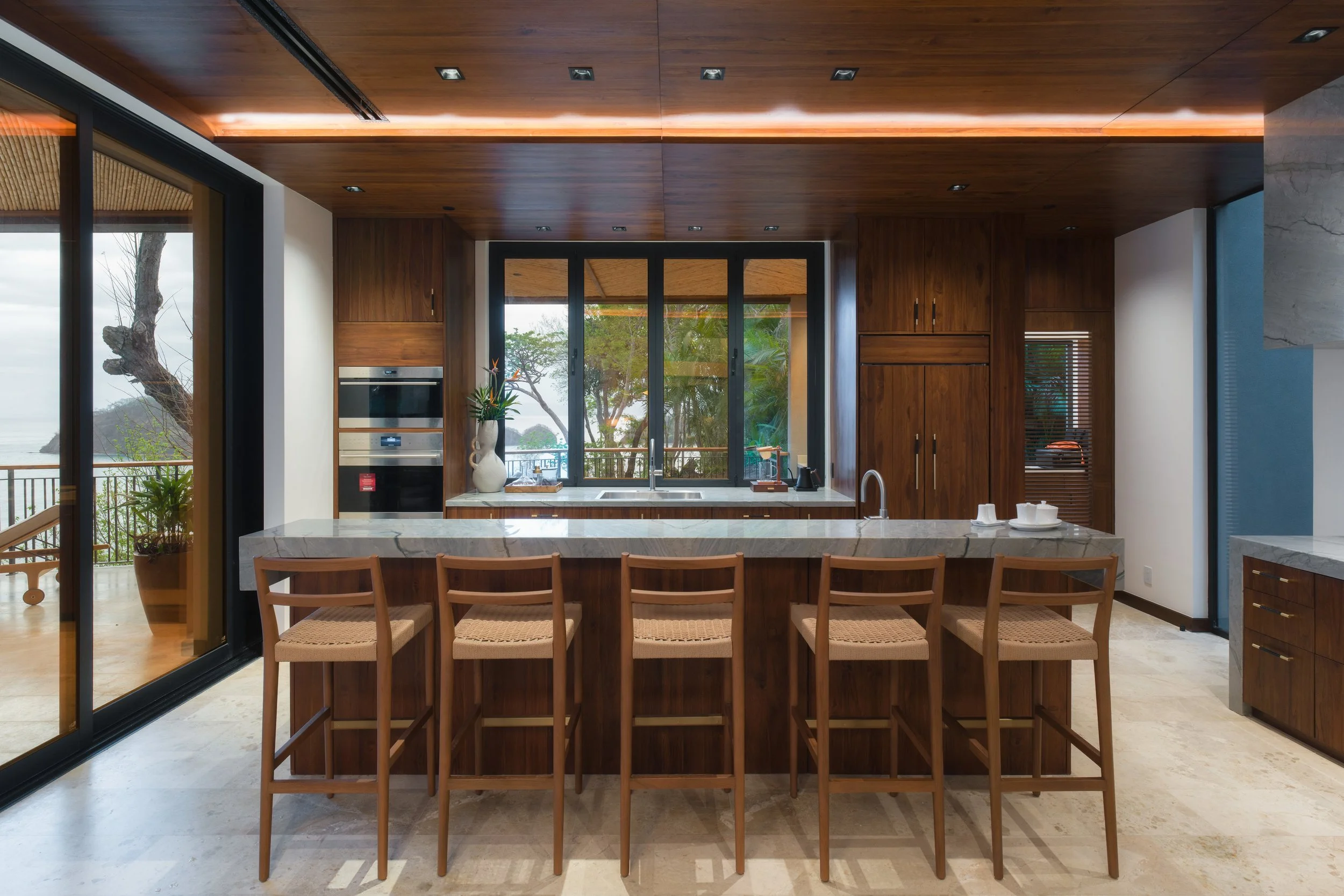 Modern kitchen with wooden cabinetry, marble countertop island with five bar stools, large window overlooking trees, kitchen appliances built into cabinets, sliding glass door leading to outdoor deck with railing.