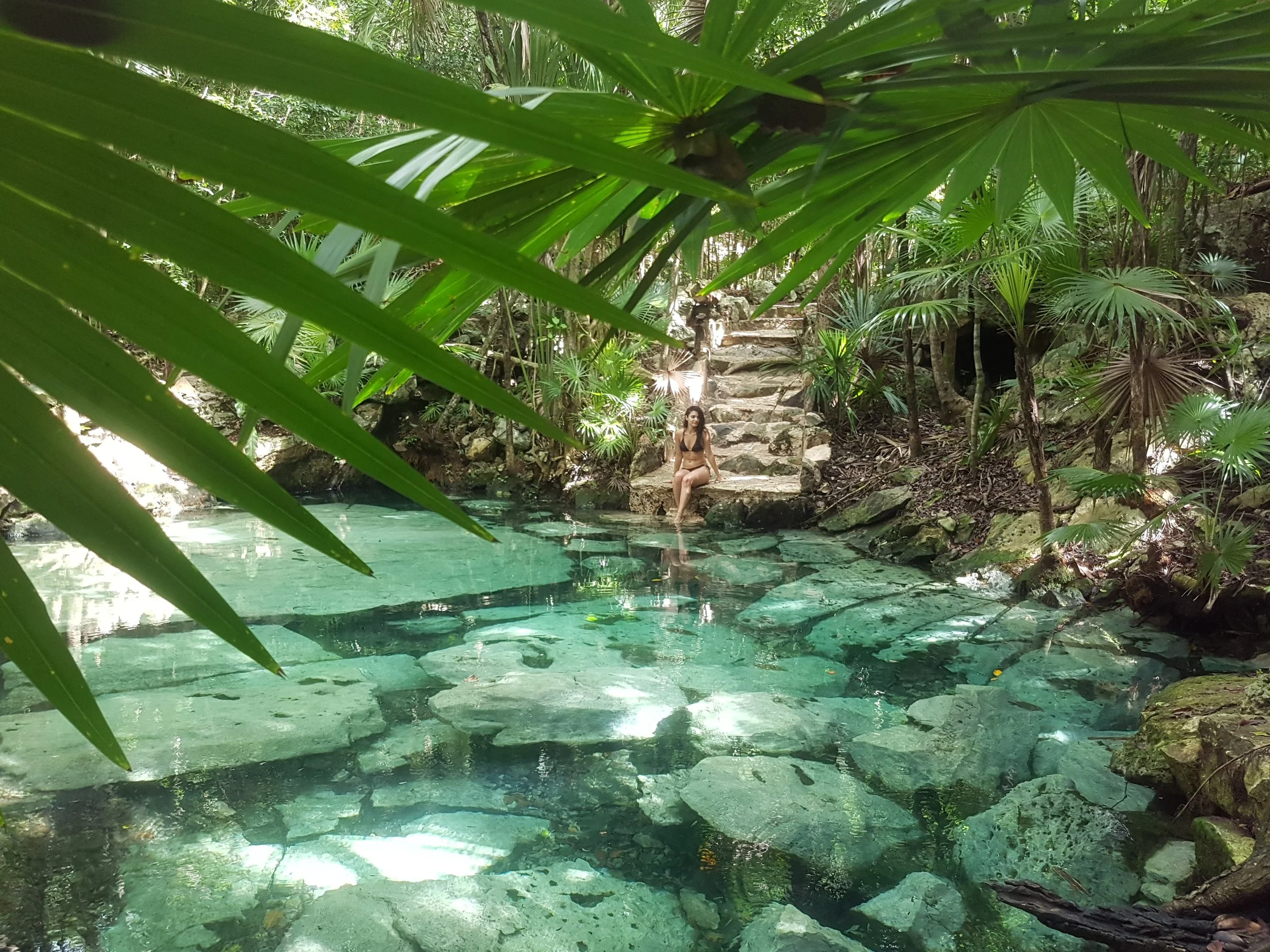 A woman sitting on rocks by a clear, shallow creek surrounded by lush green tropical plants and trees