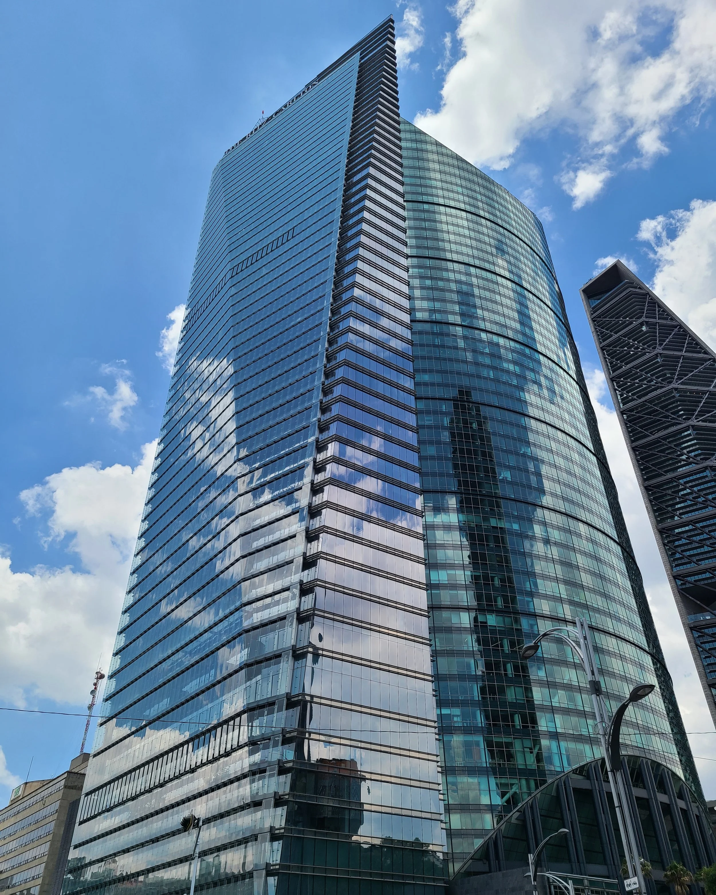 A modern glass skyscraper with reflective windows, set against a blue sky with some clouds, surrounded by other tall buildings.