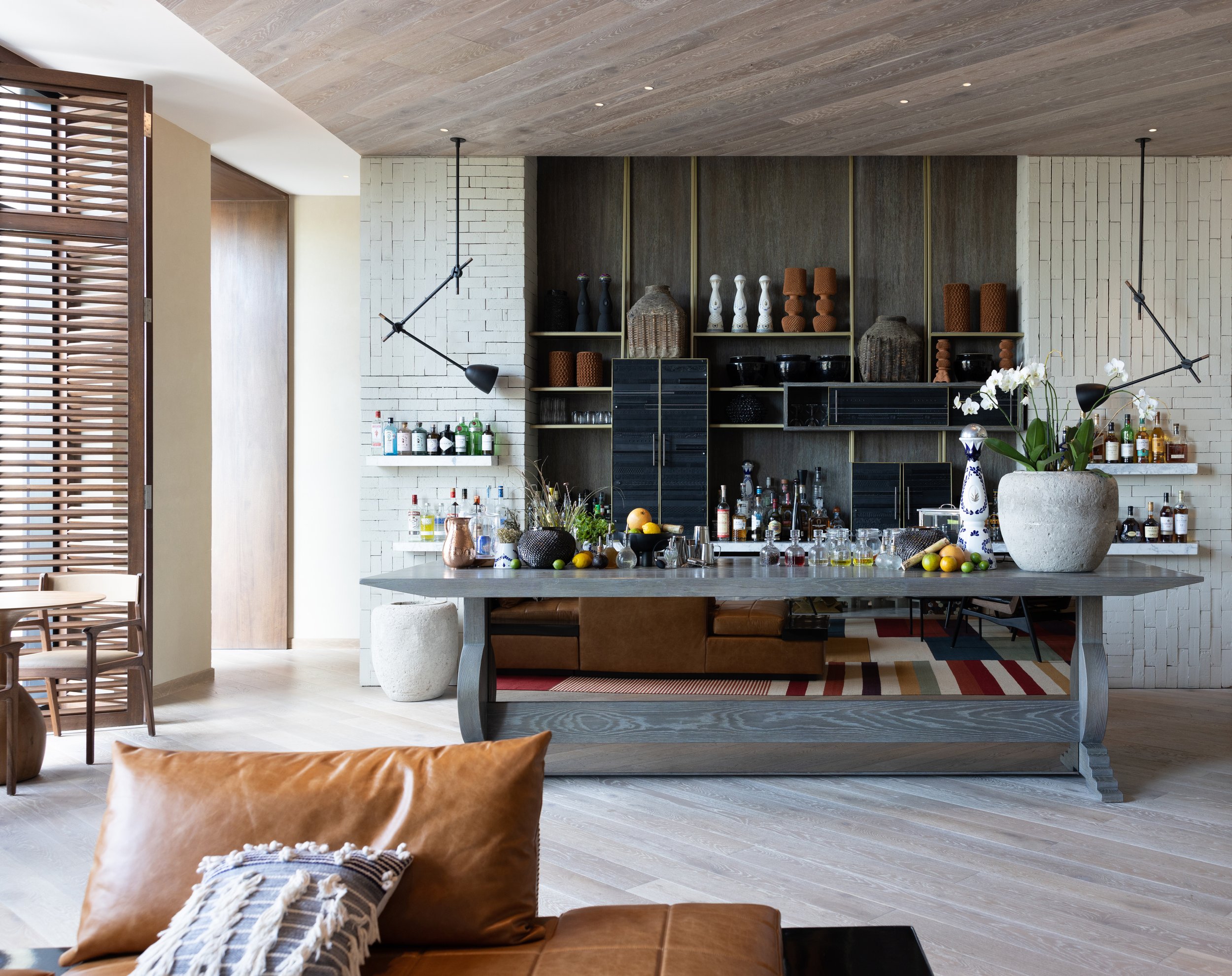 Modern living room with a bar area, wooden accents, a large table with drinks and fruit, and a leather sofa with pillows.