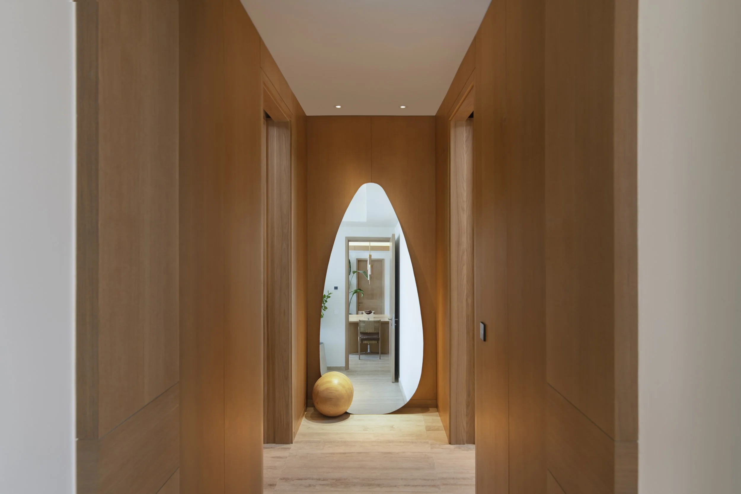 Wood-paneled hallway with a unique teardrop-shaped mirror at the end and a large wooden sphere on the floor beside it.