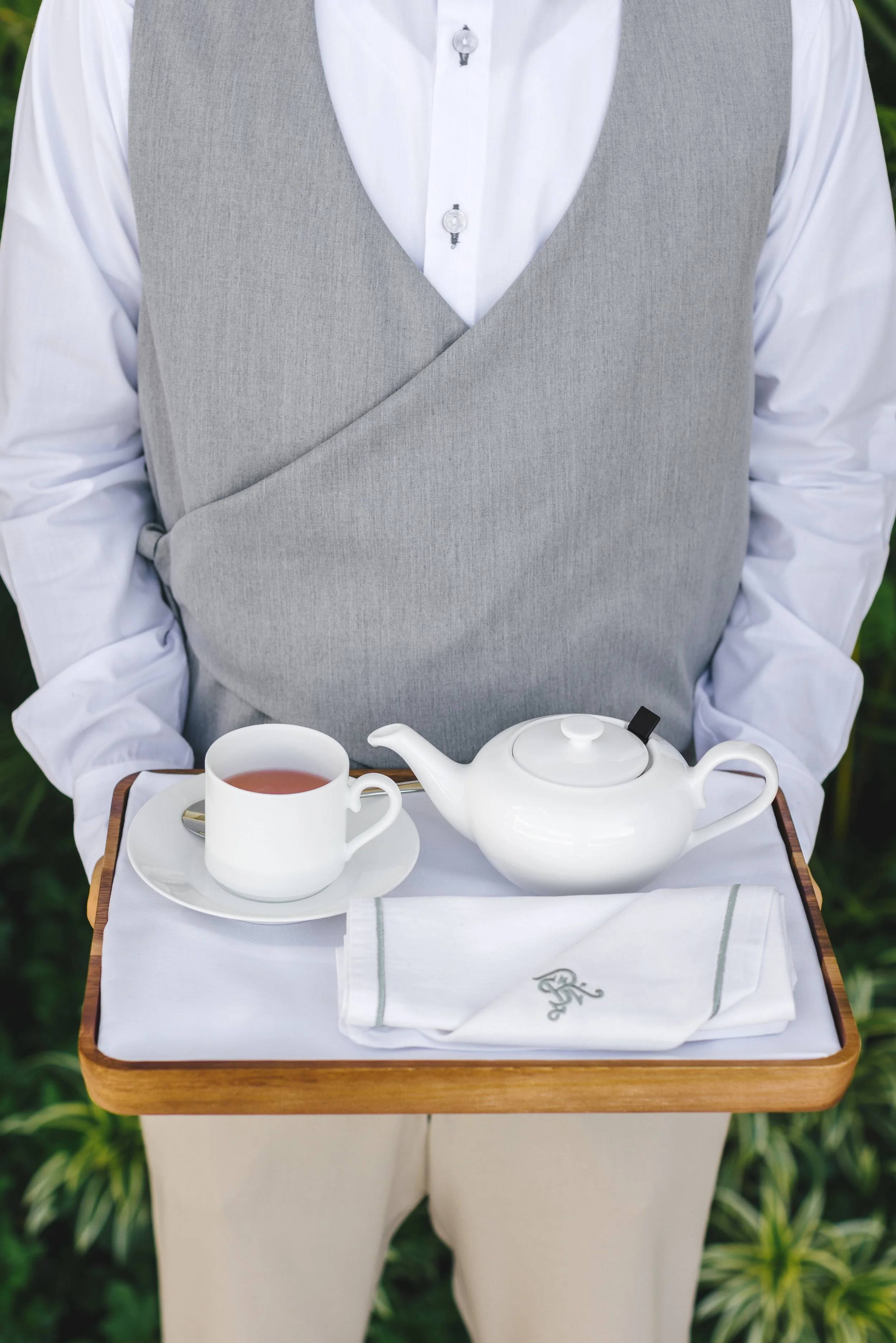 A person dressed in a white shirt and gray vest holding a wooden tray with a white teapot, a white cup with tea, and a folded white cloth napkin with an embroidered monogram.