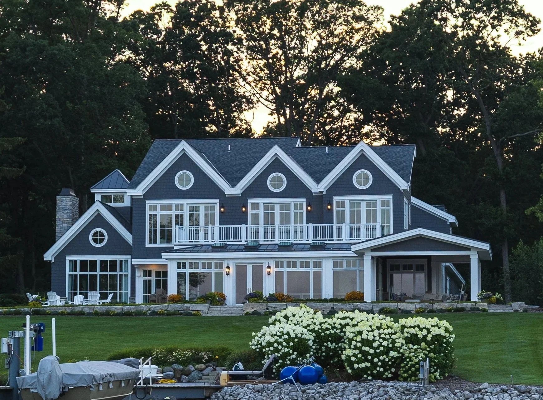 A large, modern, three-story house with blue siding and white trim, surrounded by a well-maintained lawn, with a shed or garage on the side and a small boat docked in the foreground.