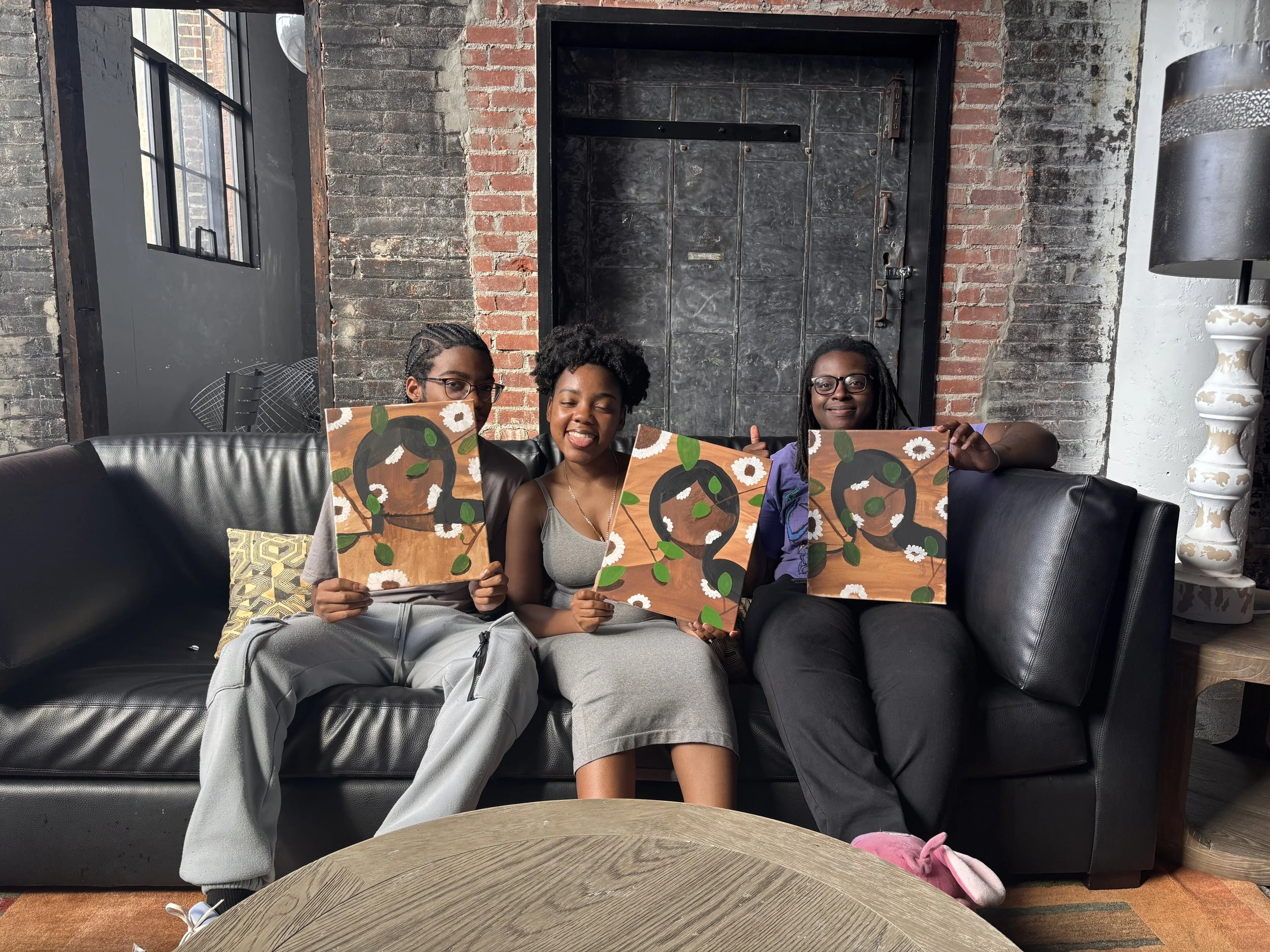 Three women sitting on a black leather sofa in an industrial-style room, holding paintings of a stylized face with green leaves and white flowers, and smiling at the camera.