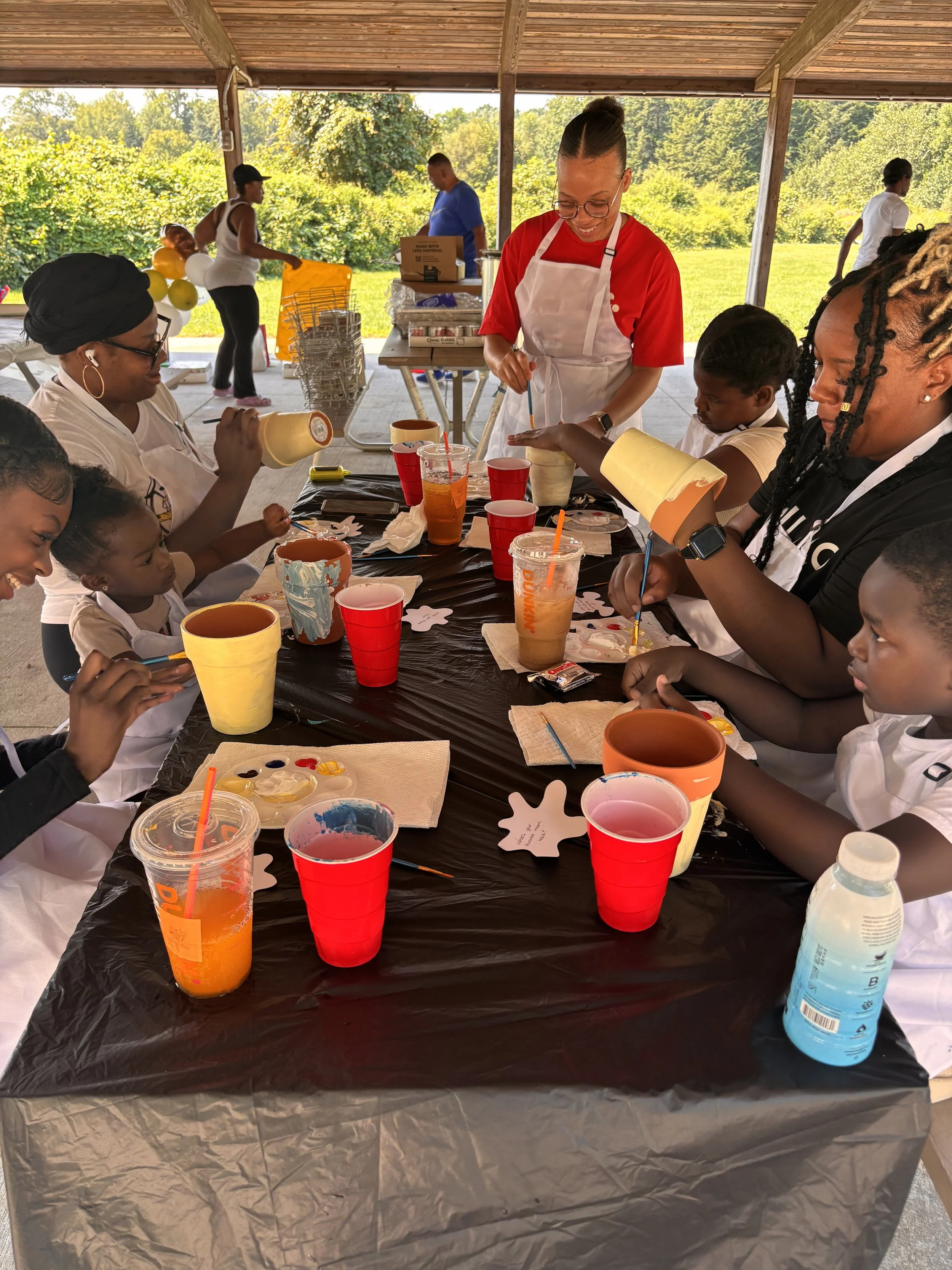 Children and adults participating in a painting activity at a gathering under a pavilion, with outdoor greenery in the background.