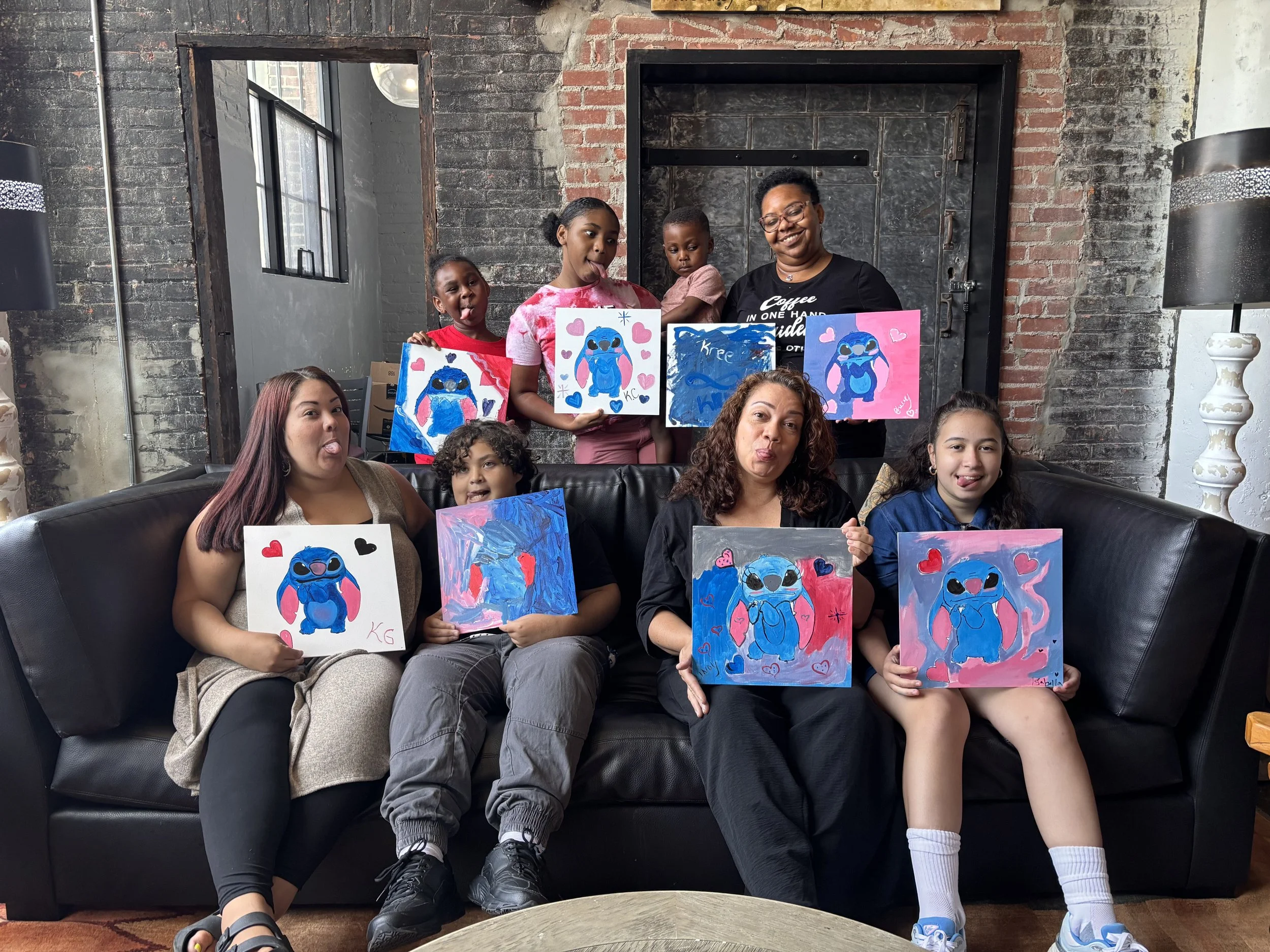 Group of children and women holding up paintings of Stitch from Lilo & Stitch, sitting on and standing behind a black leather couch in a room with brick and concrete walls.