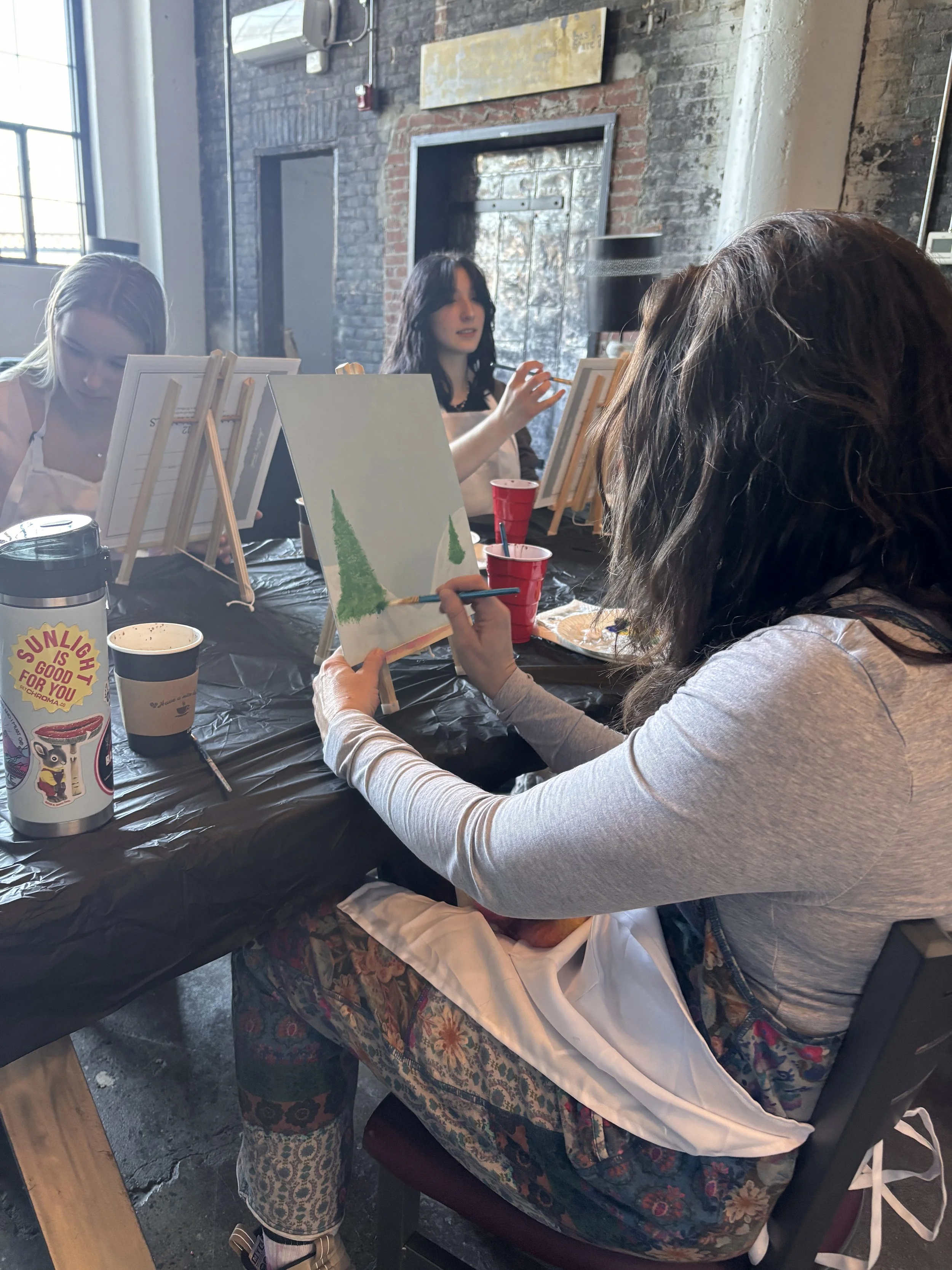 Women painting on small canvases during a group art class in a loft-style studio with brick walls and large windows.