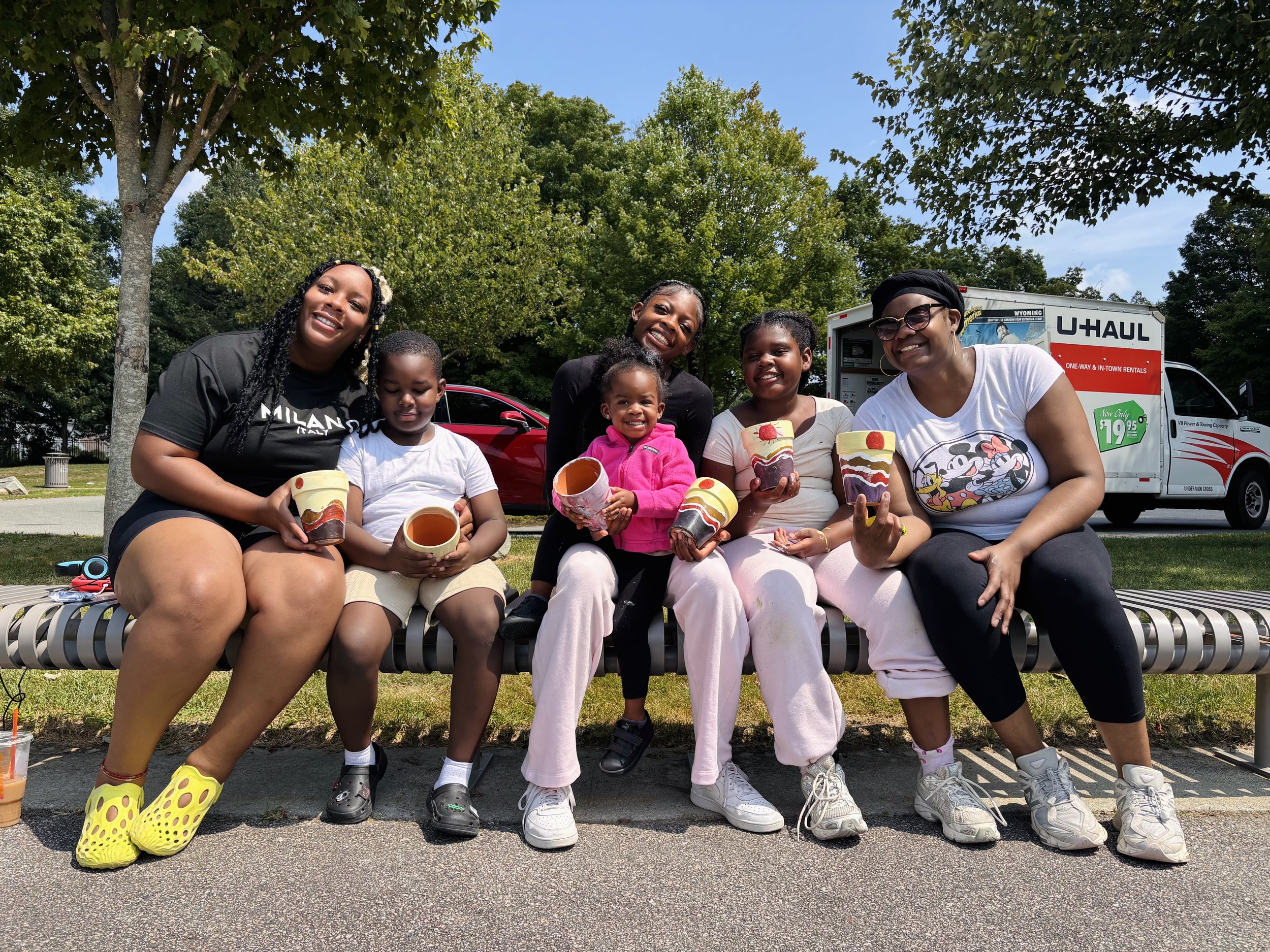 Group of six smiling people sitting on a park bench outdoors, holding colorful ice cream treats, with a U-Haul truck and cars in the background.