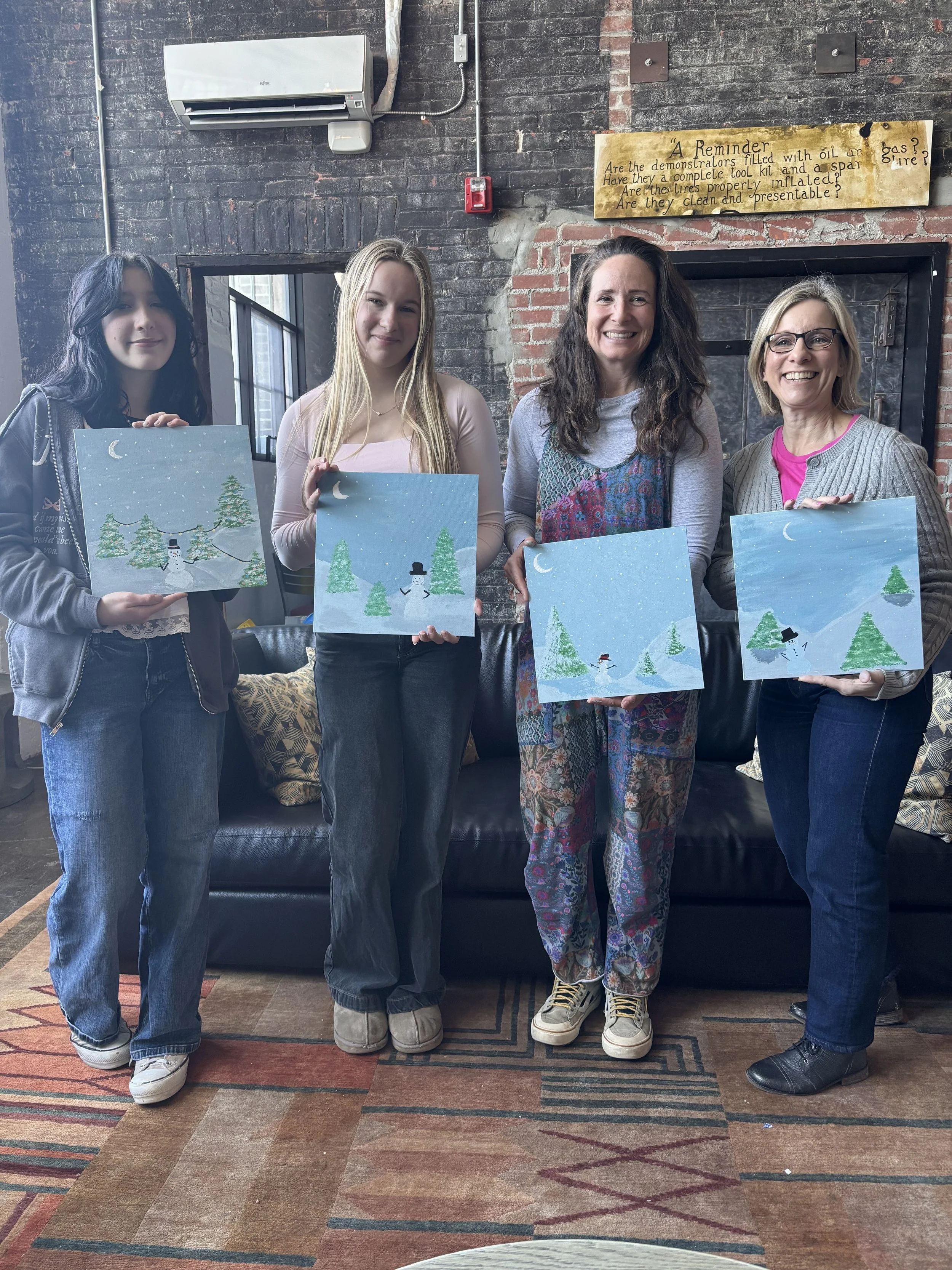 Four women and two girls are standing in front of a brick wall, each holding a painting of a winter scene with snow, trees, and a snowman. They are in a cozy indoor setting.