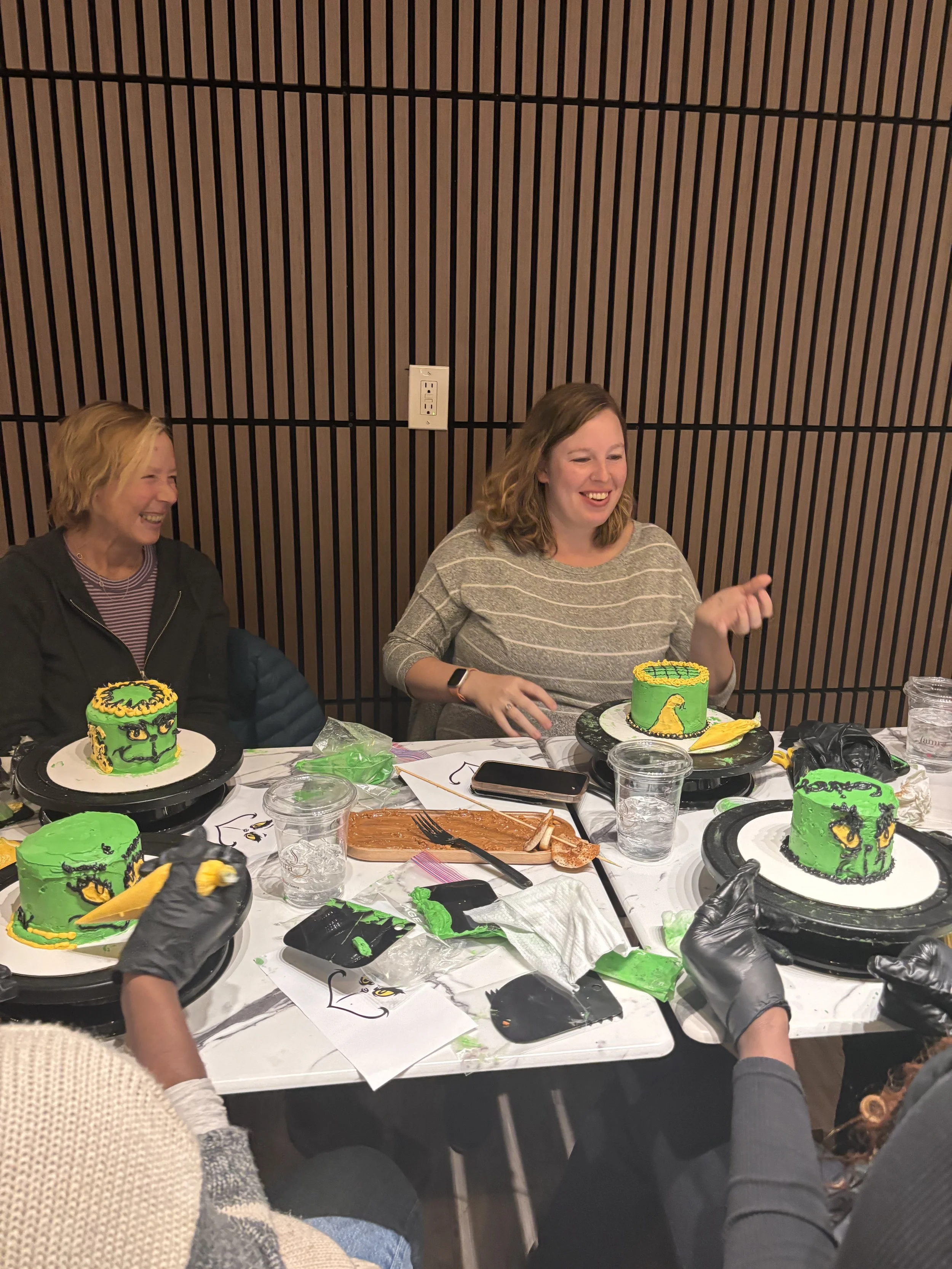People gathered around a table decorating green and yellow dragon-themed cakes, with two women smiling and laughing, some wearing black gloves, in a room with wooden panel walls.