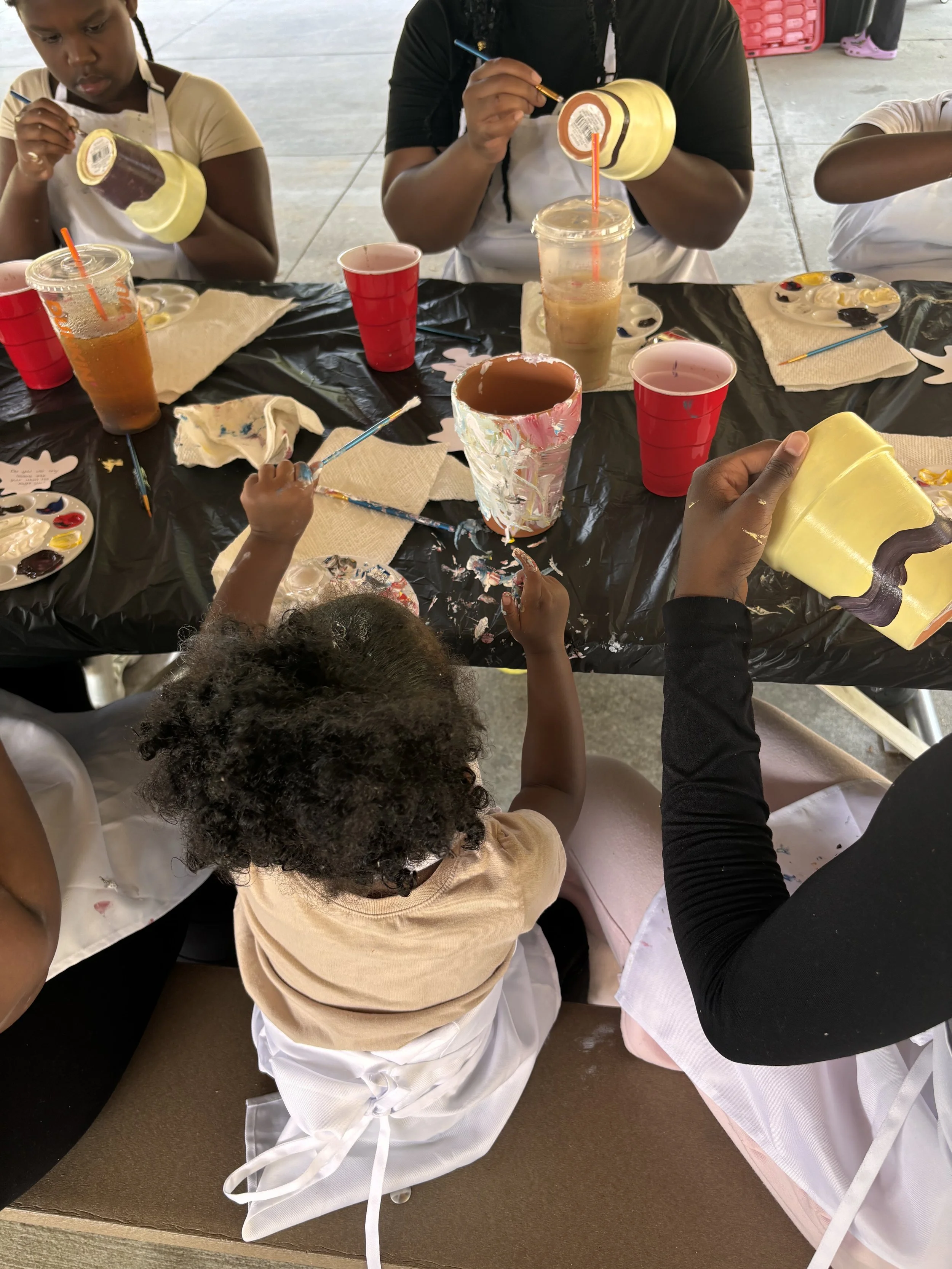 Children and adults painting and decorating ceramic mugs at a table covered with black tablecloths, paint palettes, and cups of drinks.